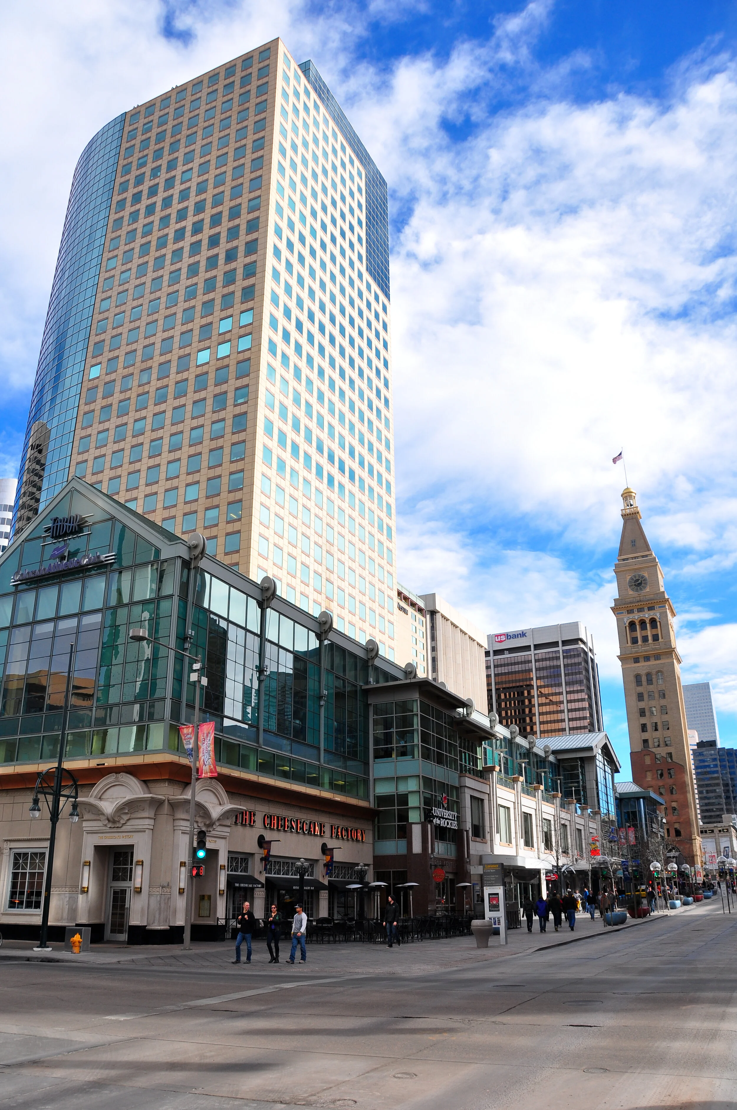 Downtown city street with modern high-rise buildings, an older clock tower, and people walking on the sidewalk. Clear blue sky with some clouds.