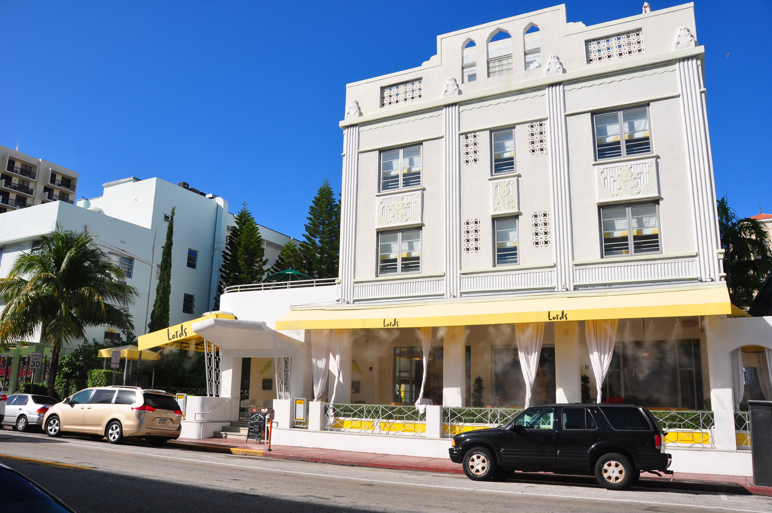 White multi-story building with Art Deco architecture, yellow awning with the word "Lords", outdoor seating area with white curtains, parked cars in front, palm trees and adjacent buildings, clear blue sky.