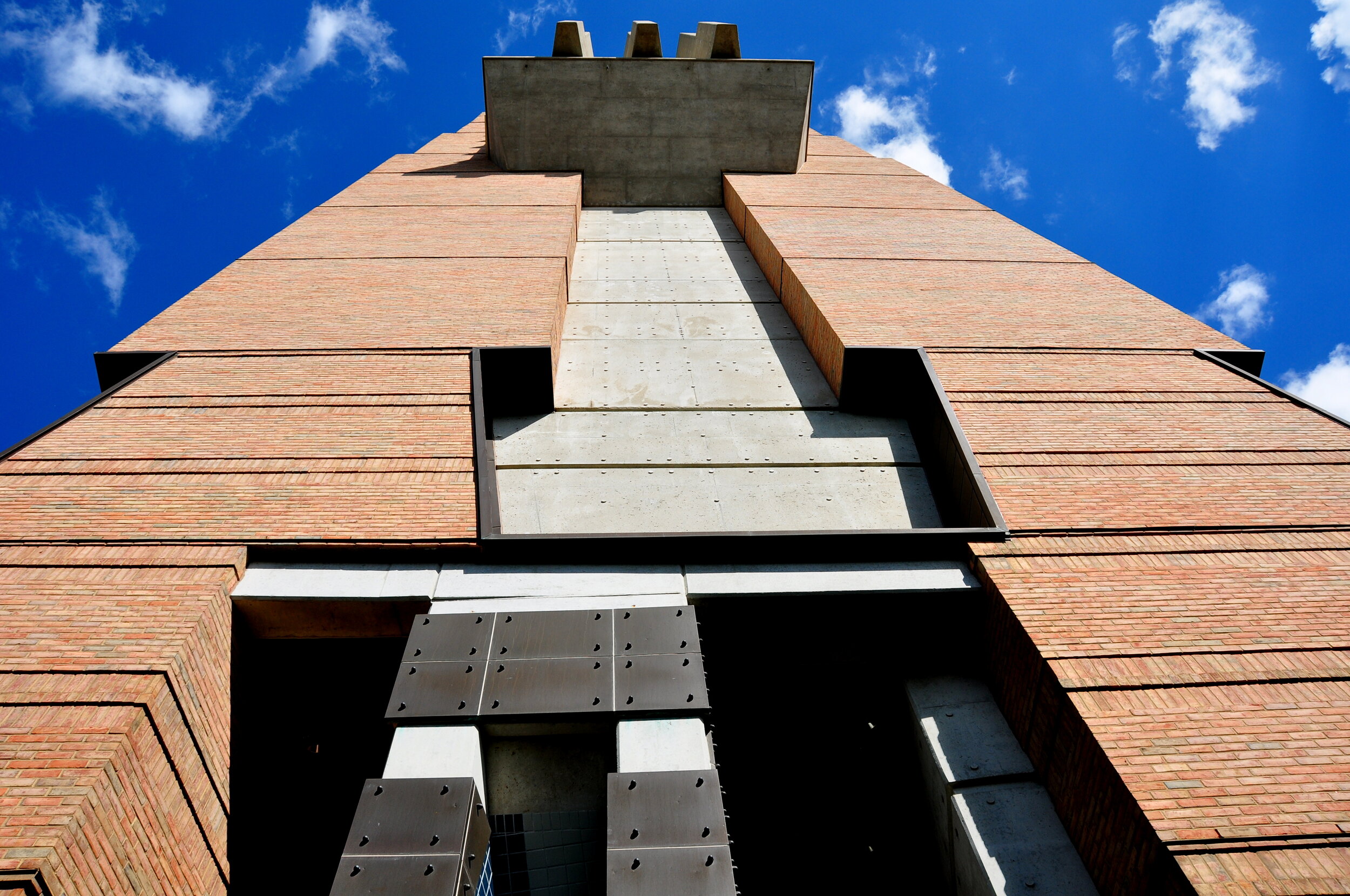 Low-angle view of a tall brick building with grey concrete sections, against a bright blue sky with scattered white clouds.