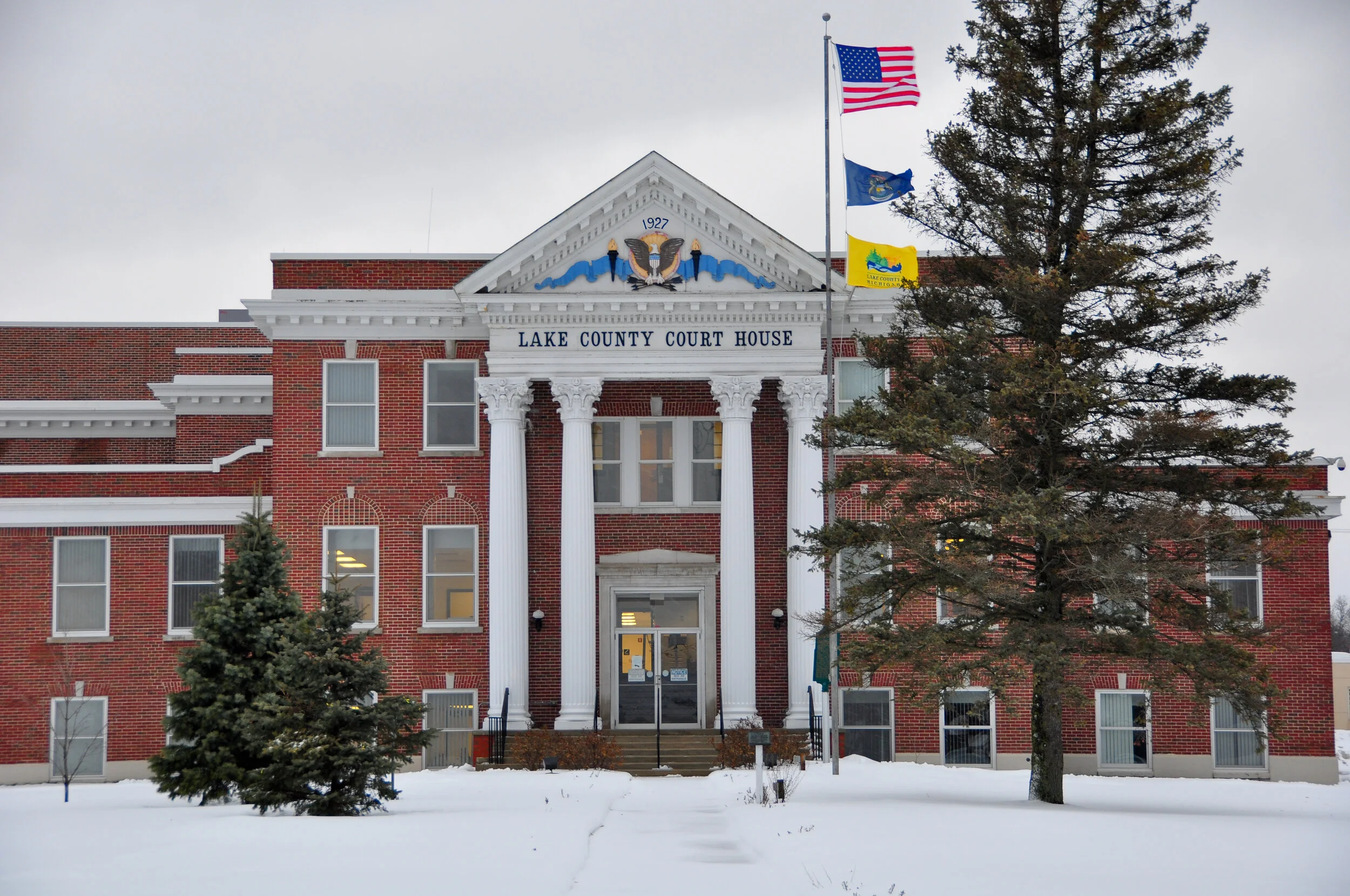 The Lake County Courthouse building made of red brick with white columns and trim, with flags flying on poles, including the American flag, in a snowy setting.