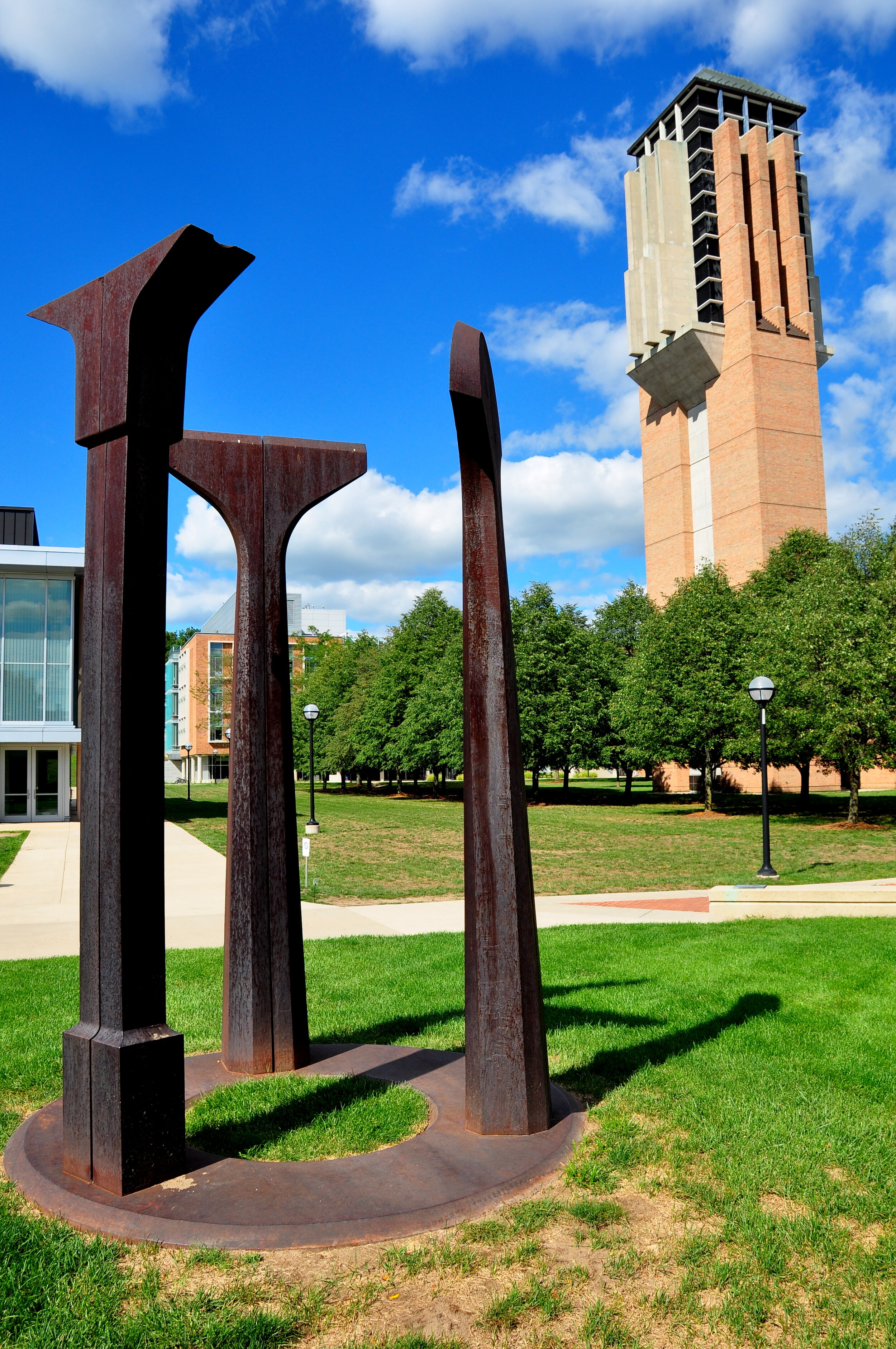 Sculpture of large, rust-colored abstract metal figures in an open park with green grass, paved walking paths, trees, and a tall brick clock tower in the background under a blue sky with white clouds.