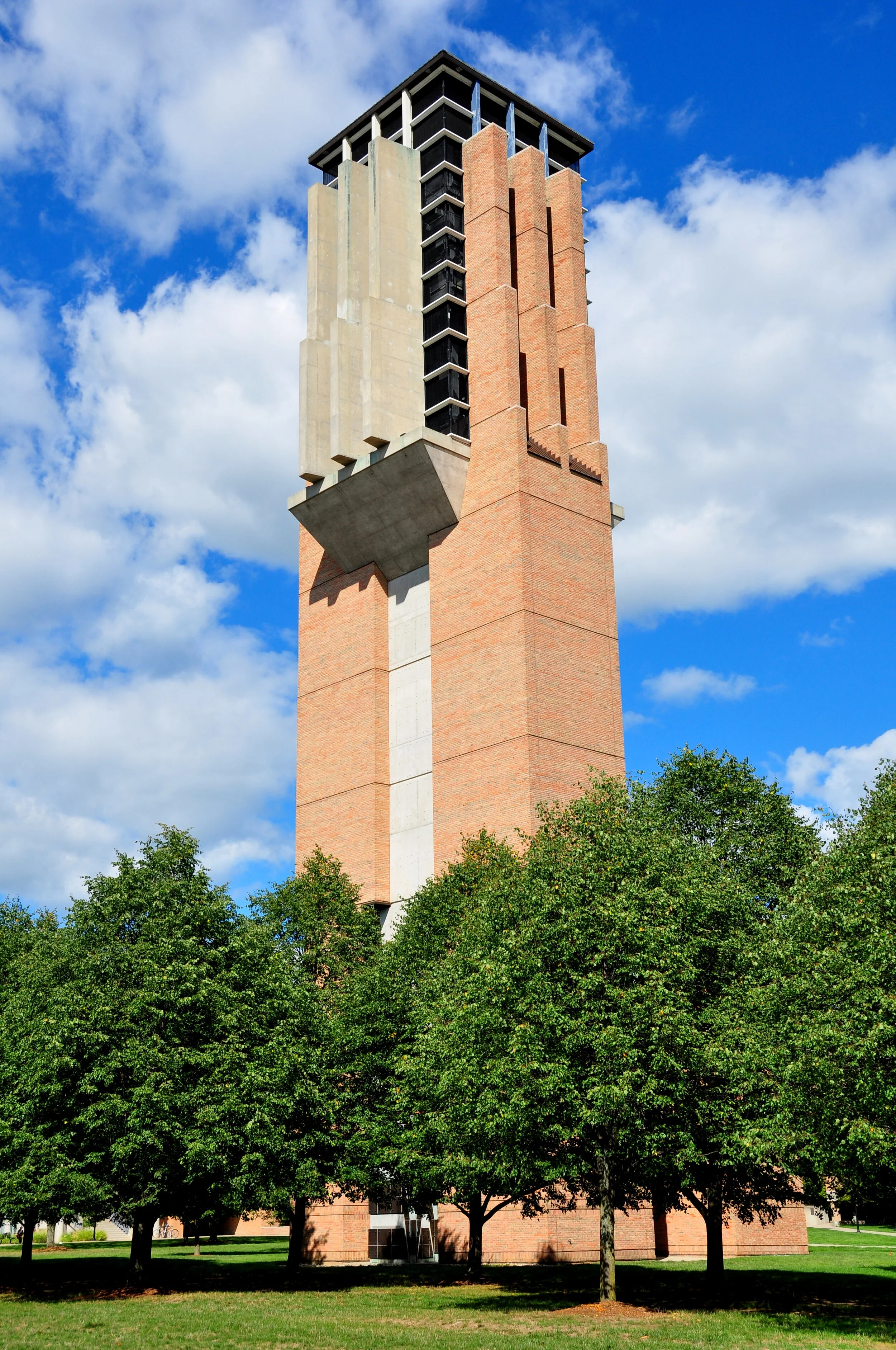 A tall, modern brick and concrete tower amidst green trees and a blue sky with scattered clouds.