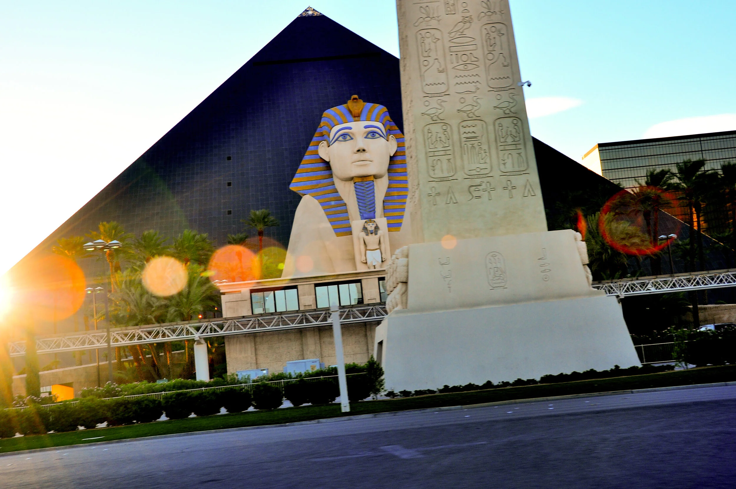 A large Egyptian-themed sculpture, including a man with a headdress, a sphinx figure, and hieroglyphic carvings, in front of a modern dark glass pyramid-shaped building with palm trees and a clear sky.