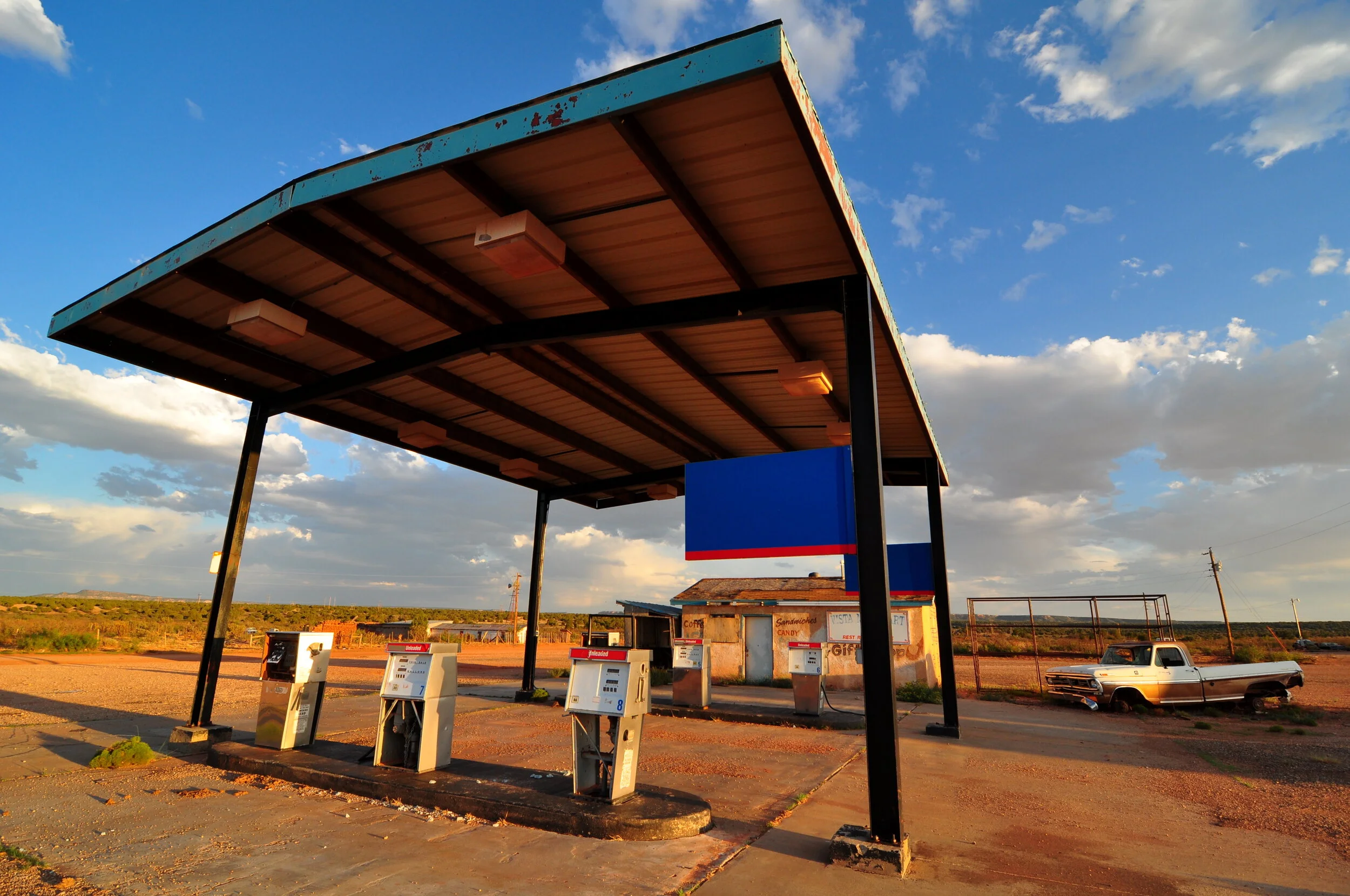 An abandoned car wash station under a large roof with four payment kiosks, a small building with signs for snacks and drinks, and a vintage pickup truck in the background on a dirt lot, under a cloudy sky during sunset.