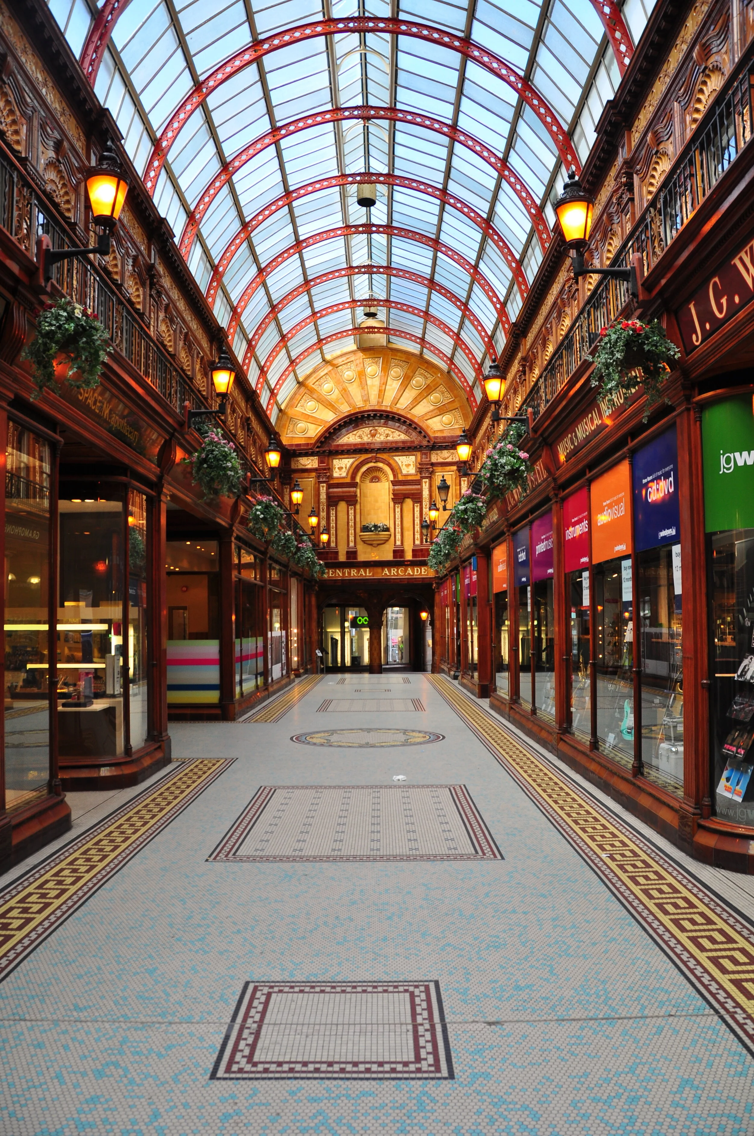 Empty shopping arcade with glass ceiling, ornate woodwork, hanging lamps, and storefronts with colorful signs.