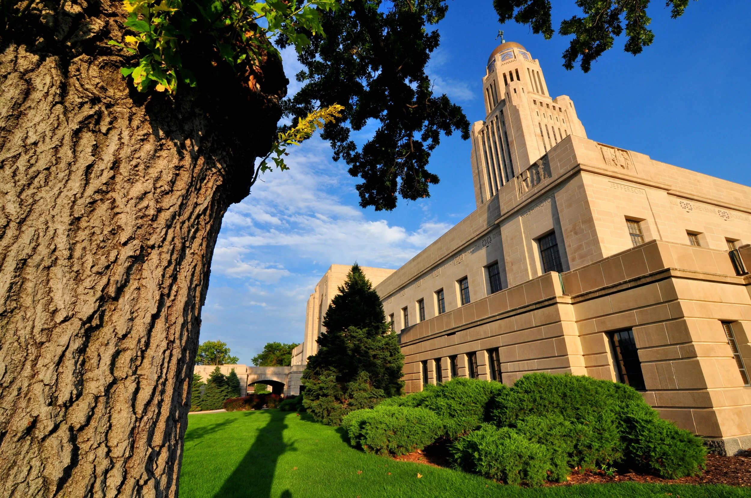 A view of a historic government building with a tall clock tower, surrounded by green trees and a well-maintained lawn, under a blue sky with some scattered clouds.