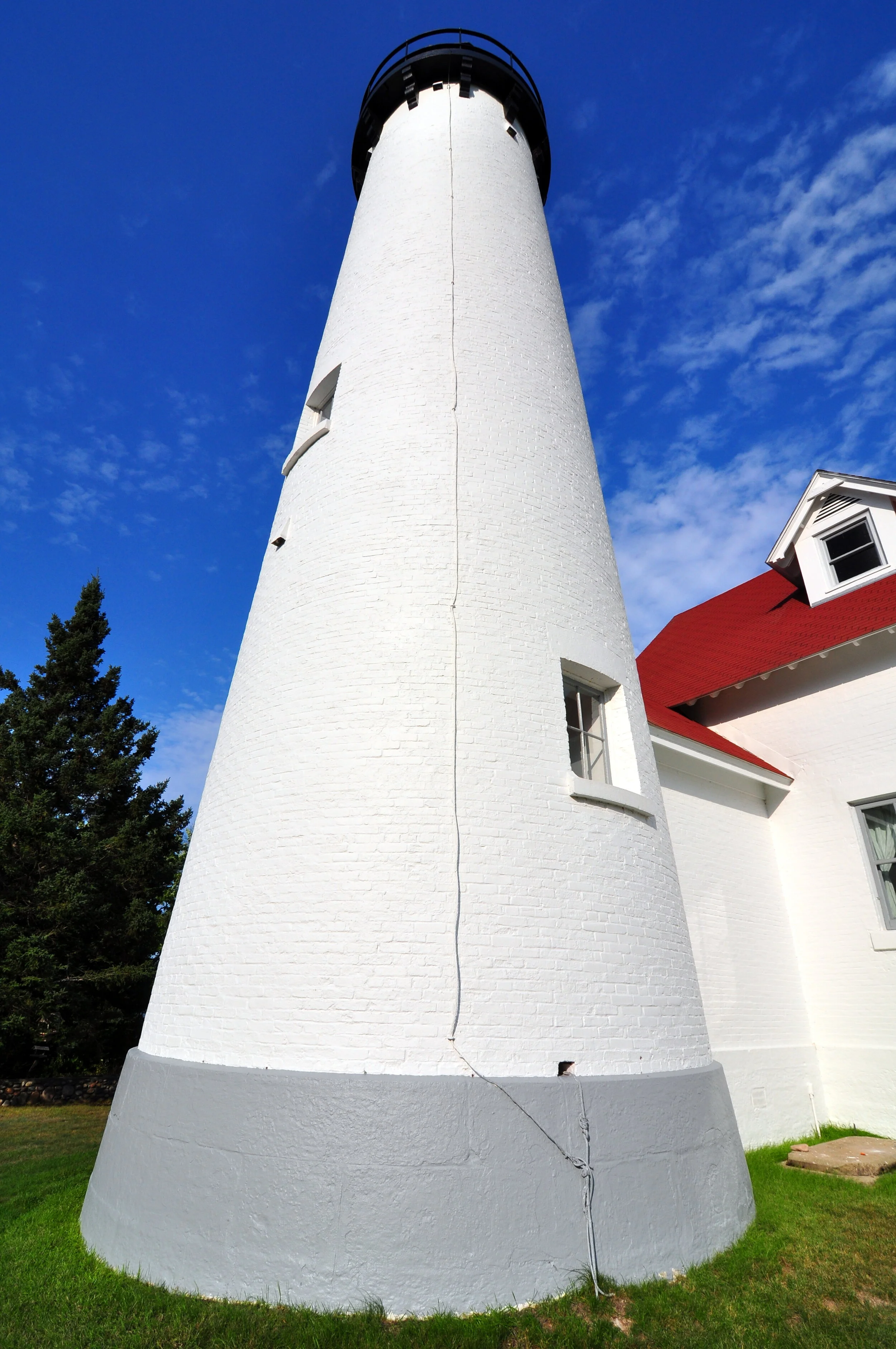 A white lighthouse with a black top and small windows, next to a white building with a red roof, under a blue sky with some clouds.