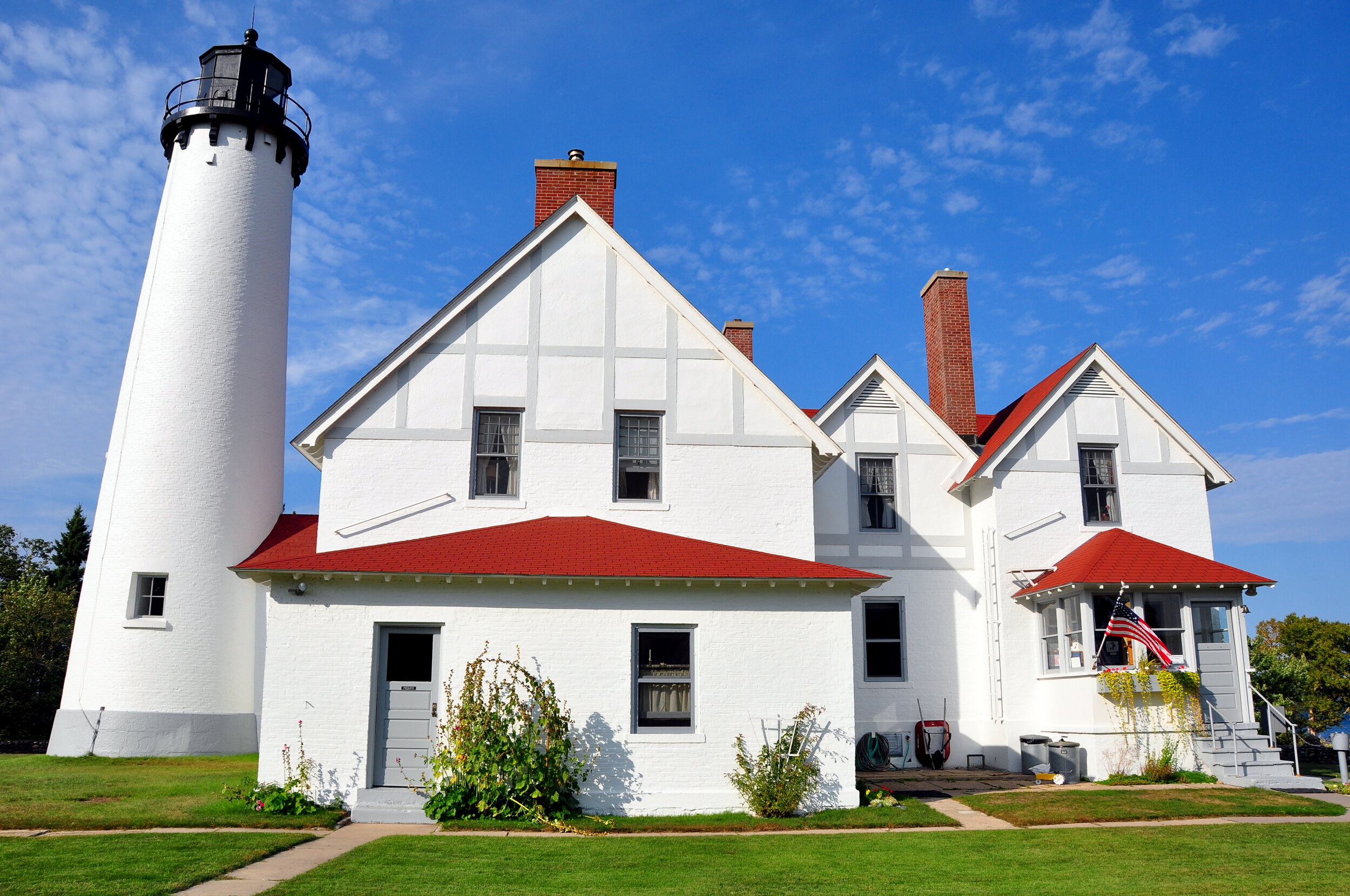 White house with a lighthouse on its left side, red roofs, and American flag, under a blue sky with scattered clouds.