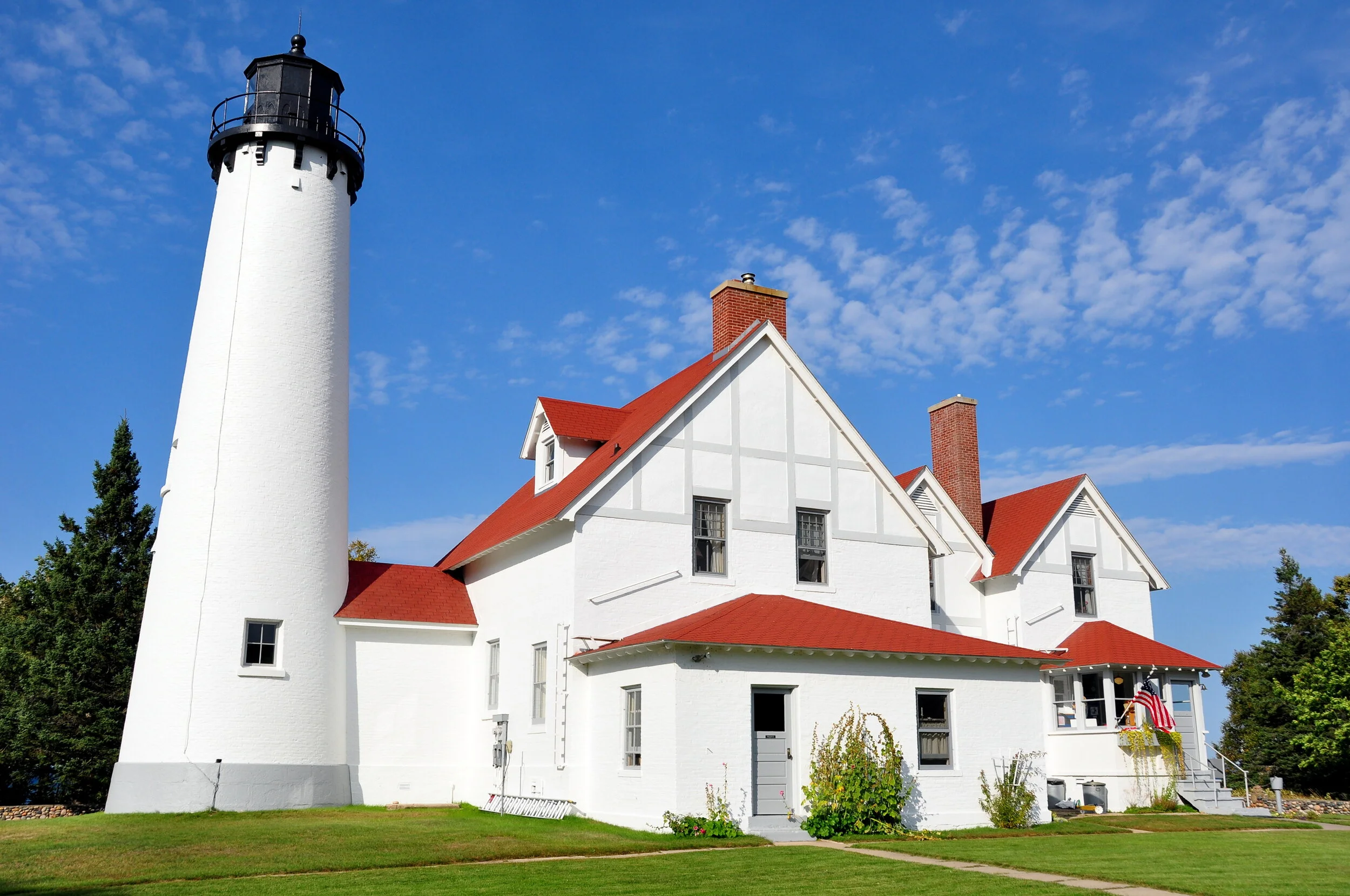 A white lighthouse with a black top next to a white house with red roof and chimneys against a blue sky with clouds.