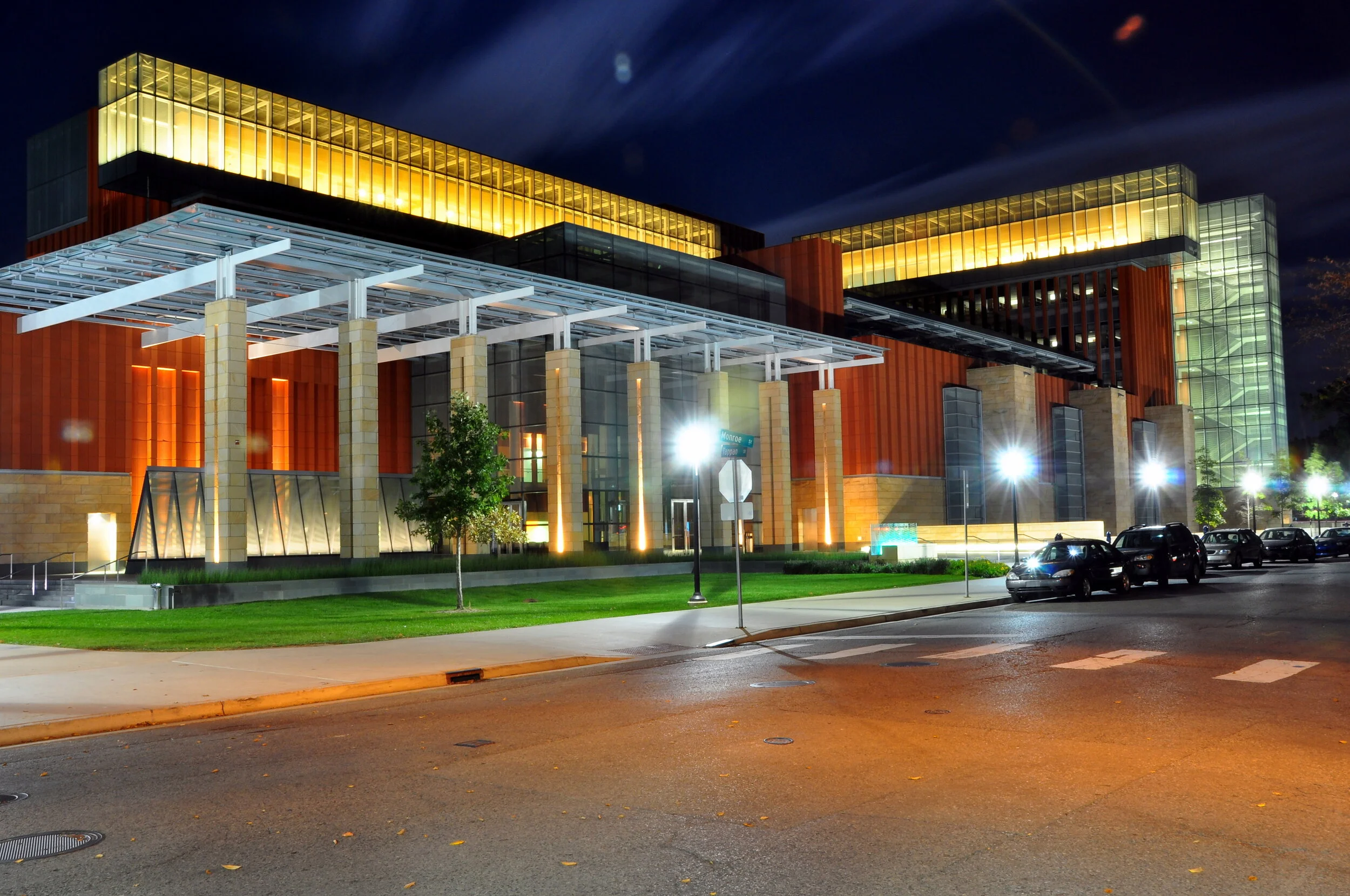 A modern, multi-story building illuminated at night, with glass and wood architectural elements, sidewalk, streetlights, and parked cars in the foreground.