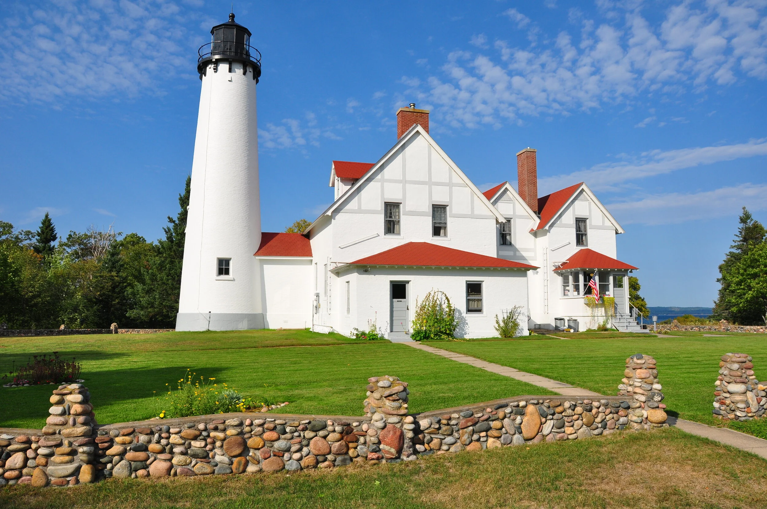 A white lighthouse with a black top and railing next to a white house with red roof and chimneys, surrounded by a green lawn, stone wall, and trees under a blue sky with clouds.