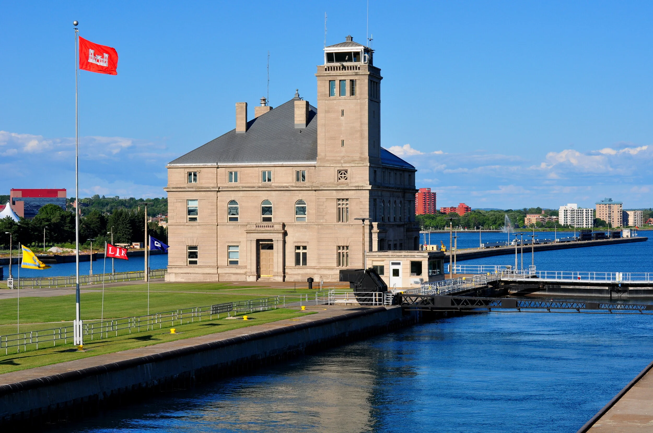 Historic stone building with a tower and multiple windows by a river, with flags on poles and modern buildings in the background under a blue sky.