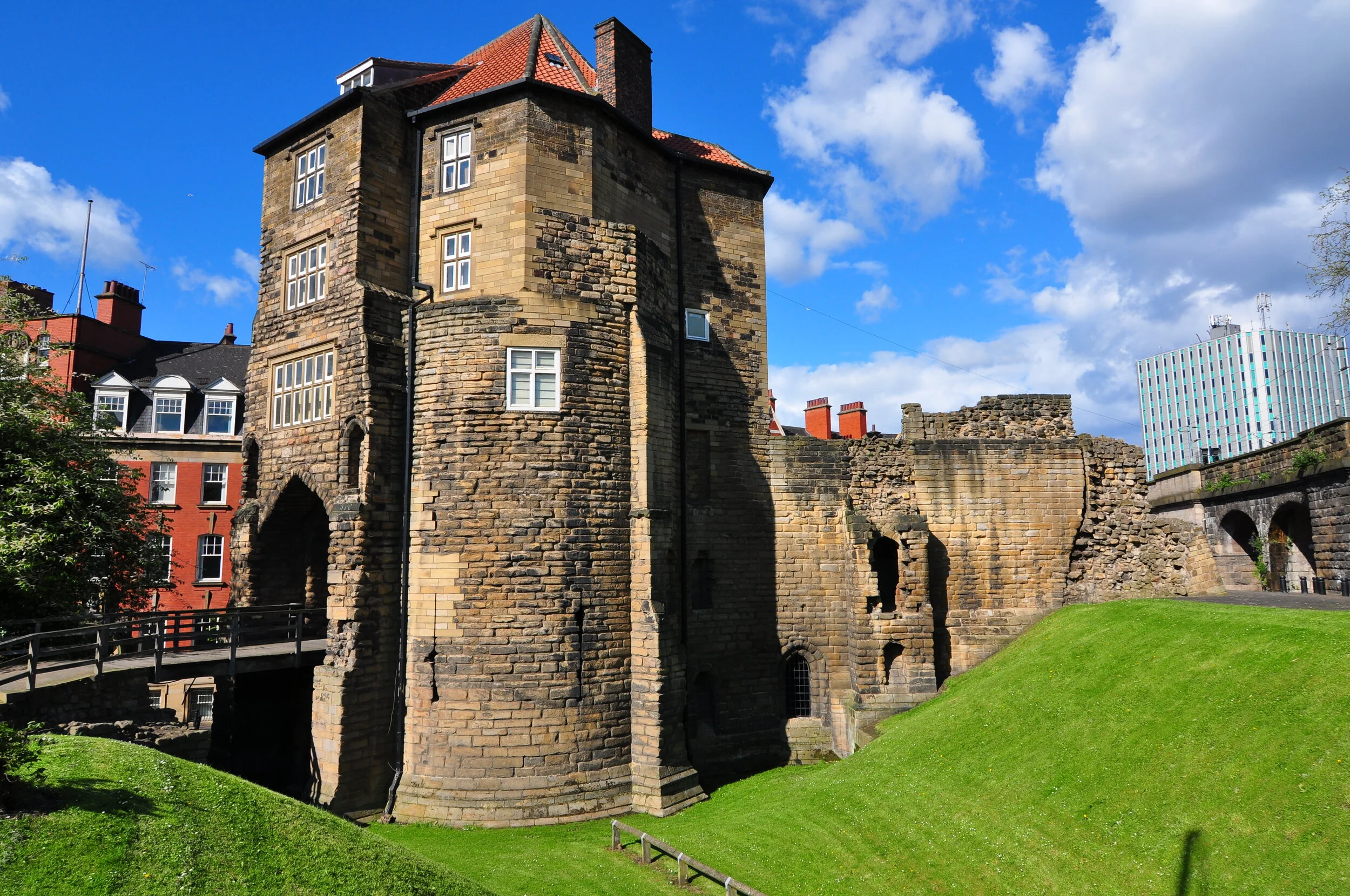 A historic stone castle with tall cylindrical towers, small windows, and a red-tiled roof, surrounded by green grass and modern buildings under a blue sky with clouds.