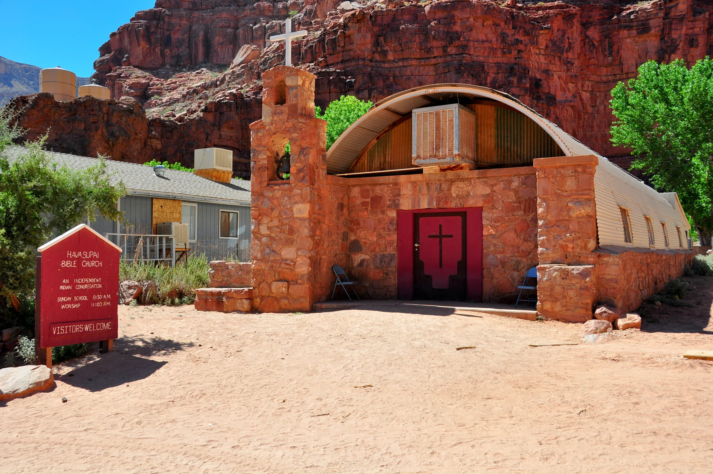 Small red rock church with a cross on the hill behind it, a red sign with white text in front, and two chairs outside the entrance, surrounded by green trees and desert landscape.