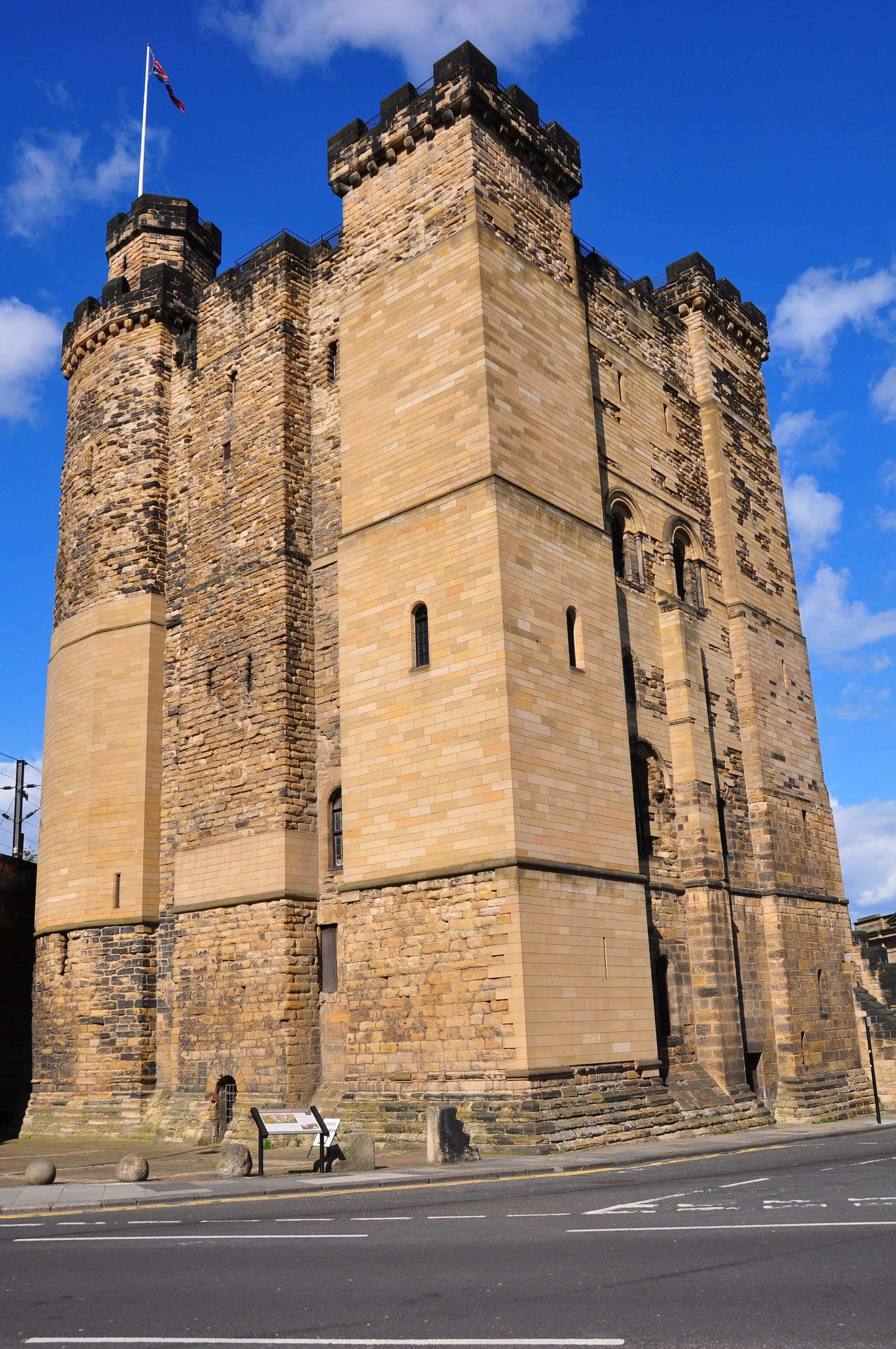 A historic stone castle with four tall cylindrical towers, featuring battlements, and a flag flying on one of the towers, set against a bright blue sky with some clouds.