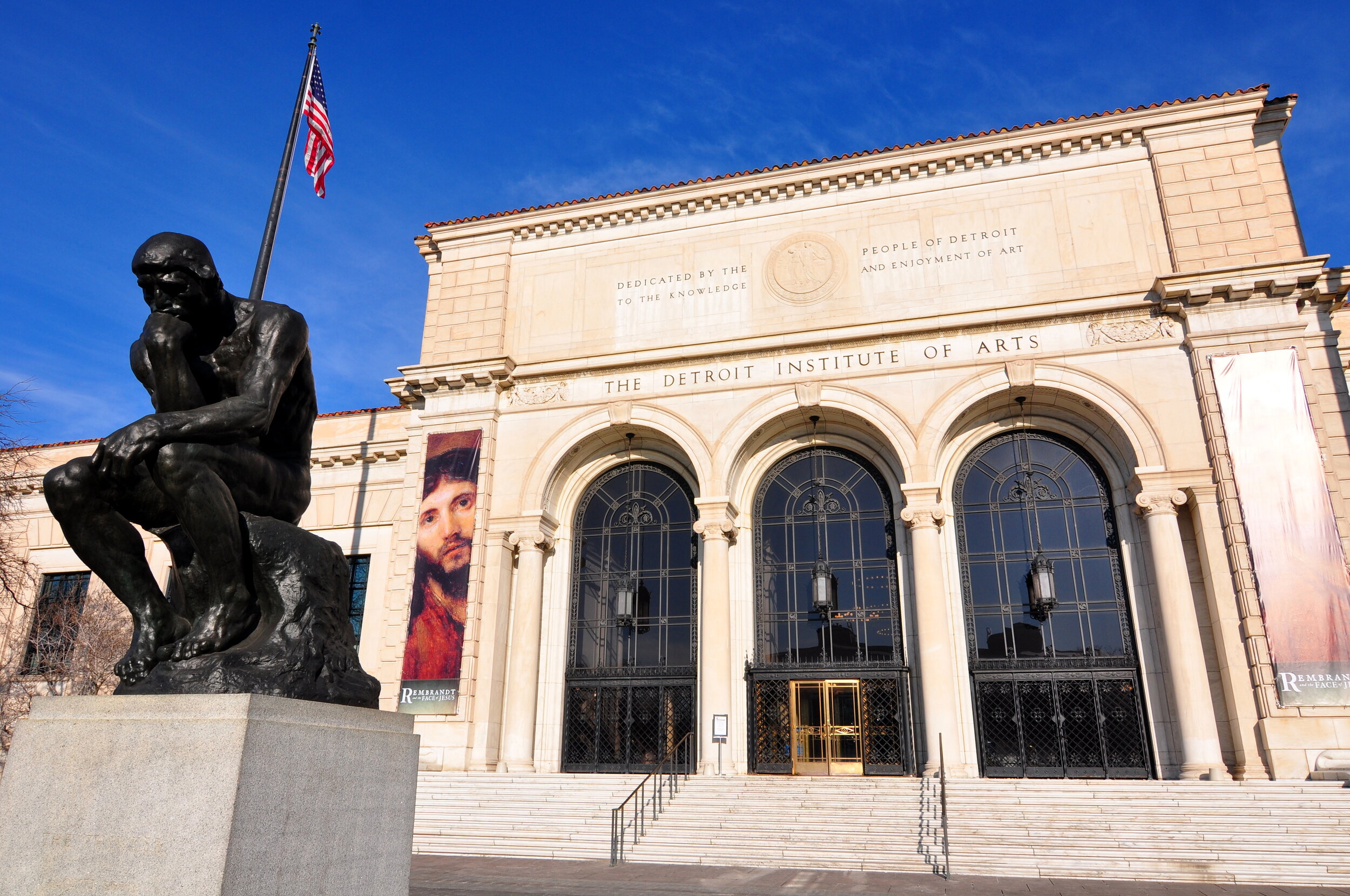 The Detroit Institute of Arts building with a statue of a thinker sculpture in front, an American flag, and a portrait of Jesus on a banner.