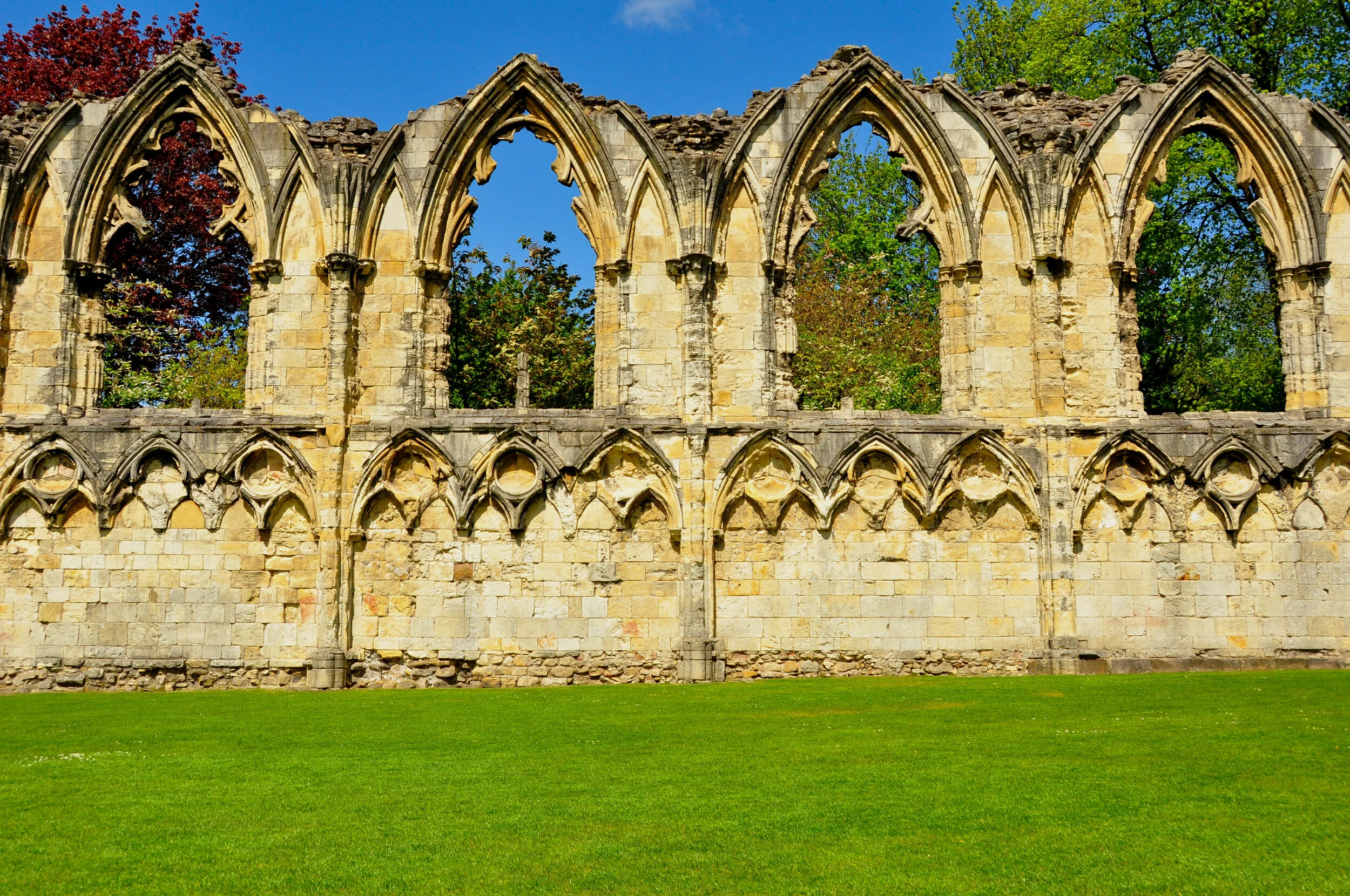 Ruins of an ancient stone building with tall, pointed arches and windows, set against a backdrop of trees with green and reddish leaves, on a sunny day with a clear blue sky, and a well-maintained grassy area in the foreground.