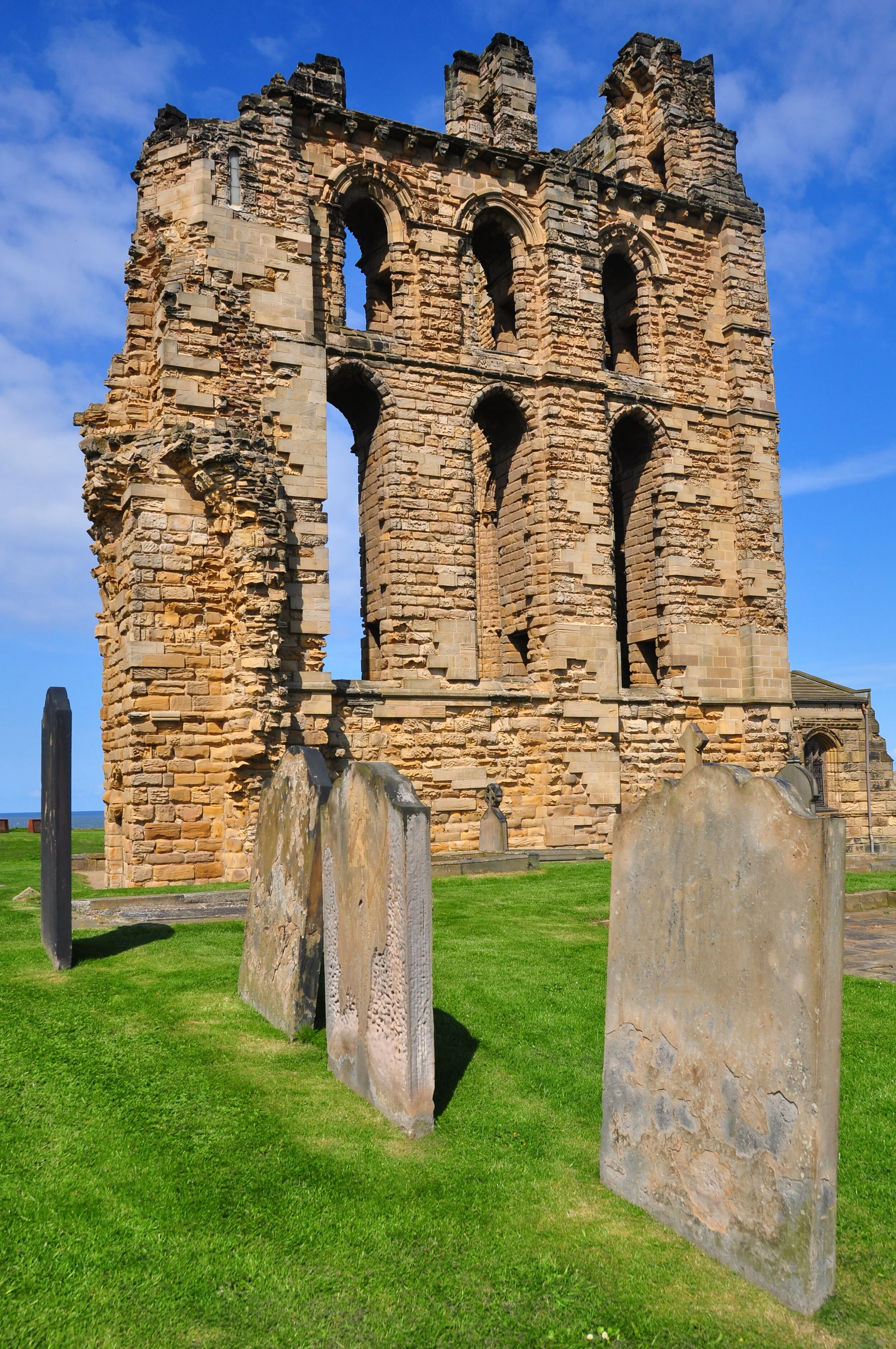 Ruins of an old stone building with six arched windows and a partially collapsed wall. Several tombstones are in the foreground on a grassy area under a blue sky with clouds.