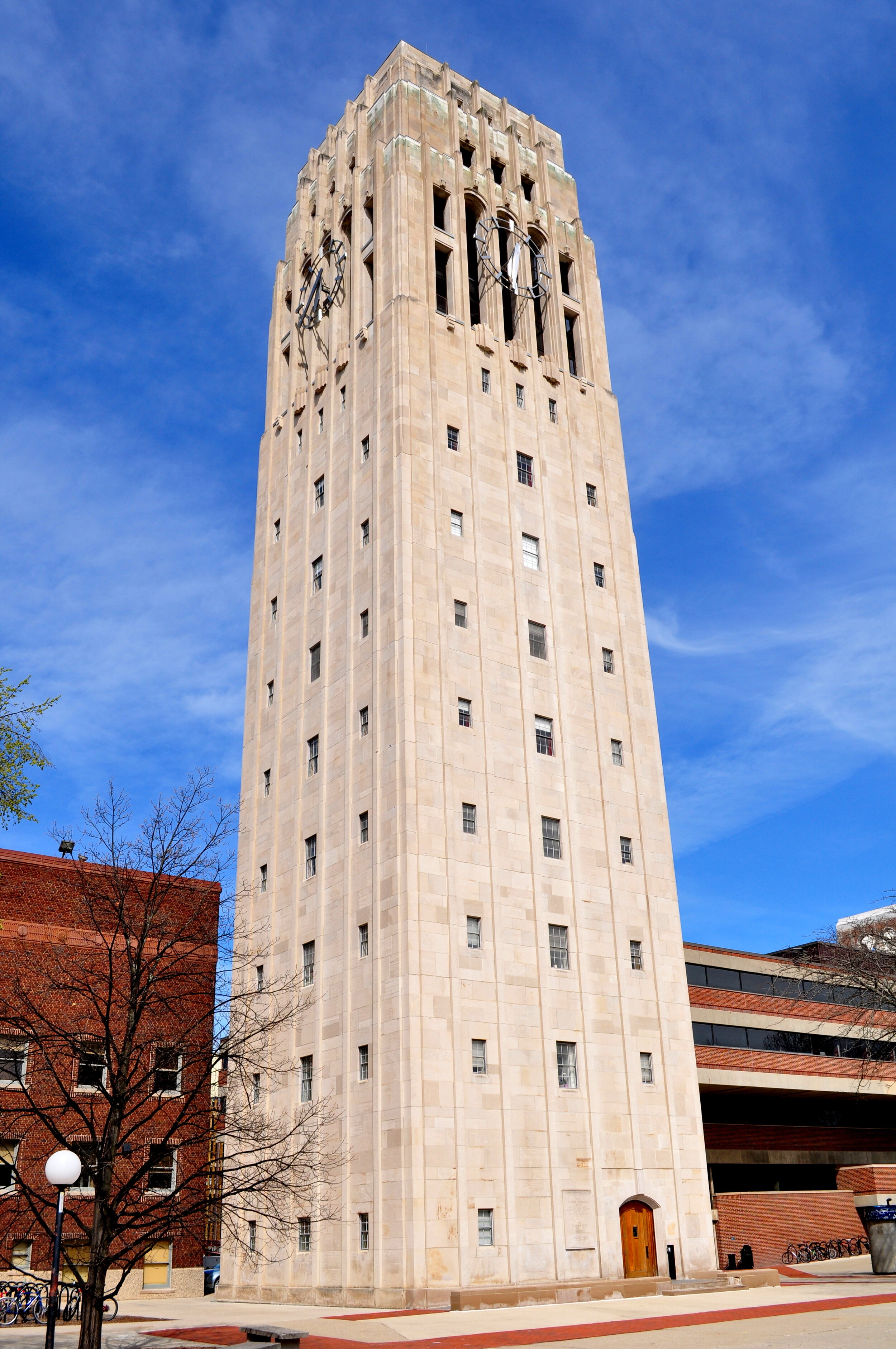 A tall stone clock tower with large clock faces near the top, set against a clear blue sky, with some trees and buildings at its base.
