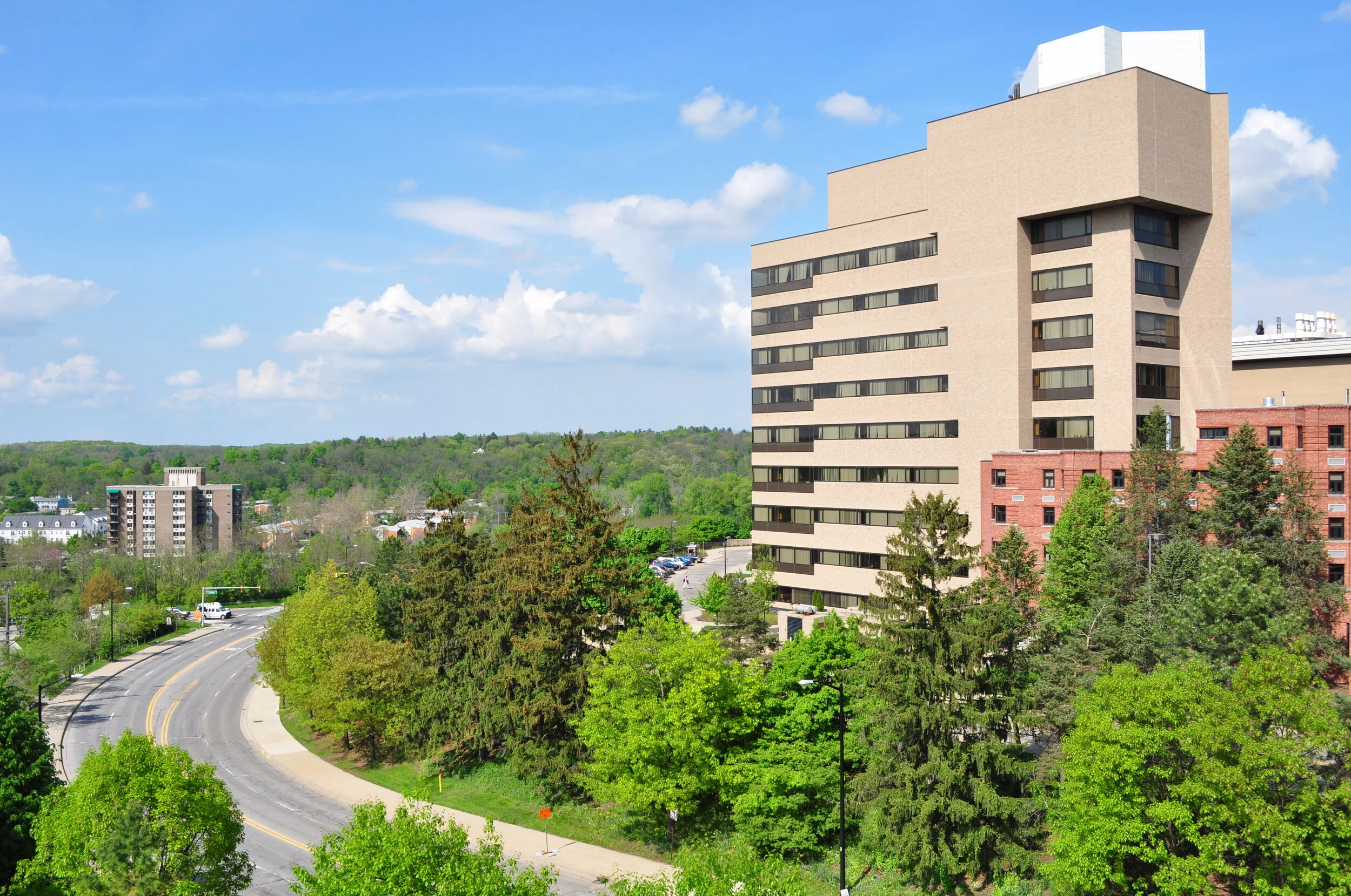 A tall beige and brown building with several horizontal windows, surrounded by lush green trees, with a winding road and a parking lot nearby, under a blue sky with clouds.