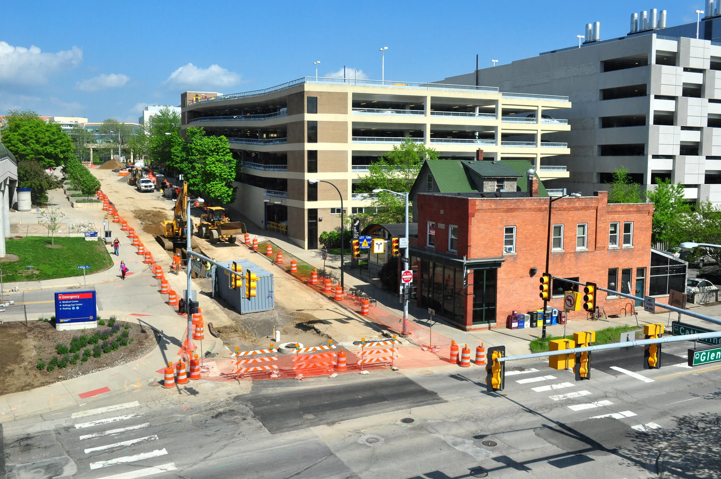 Street under construction with orange cones and barriers, a forklift, and pedestrians walking on the sidewalk. Multi-story parking garage and small brick building in the background, traffic lights, and street signs at the intersection.