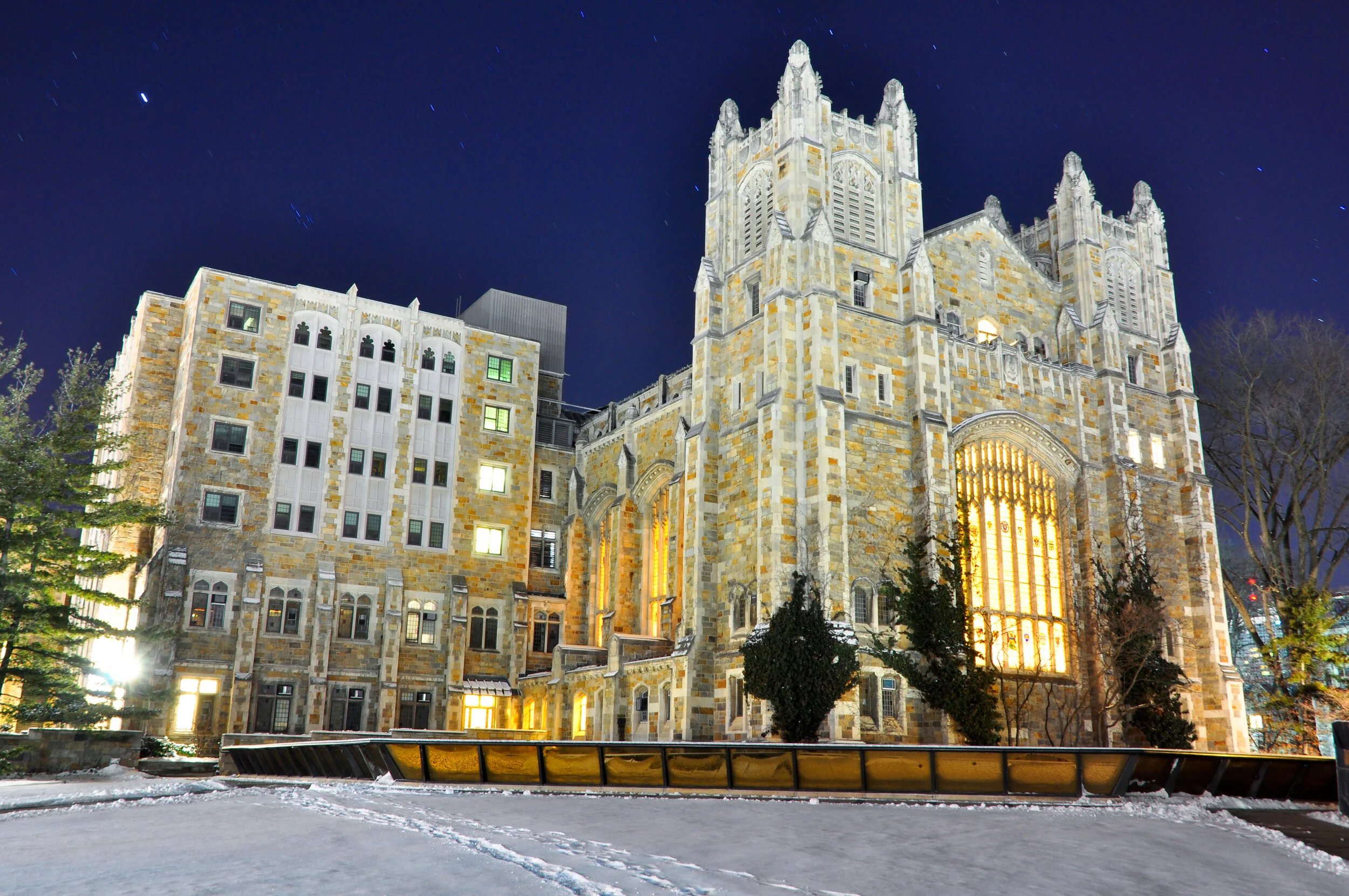 Night view of a large, illuminated stone church with gothic architecture, pointed arches, and tall windows, surrounded by snow and trees.