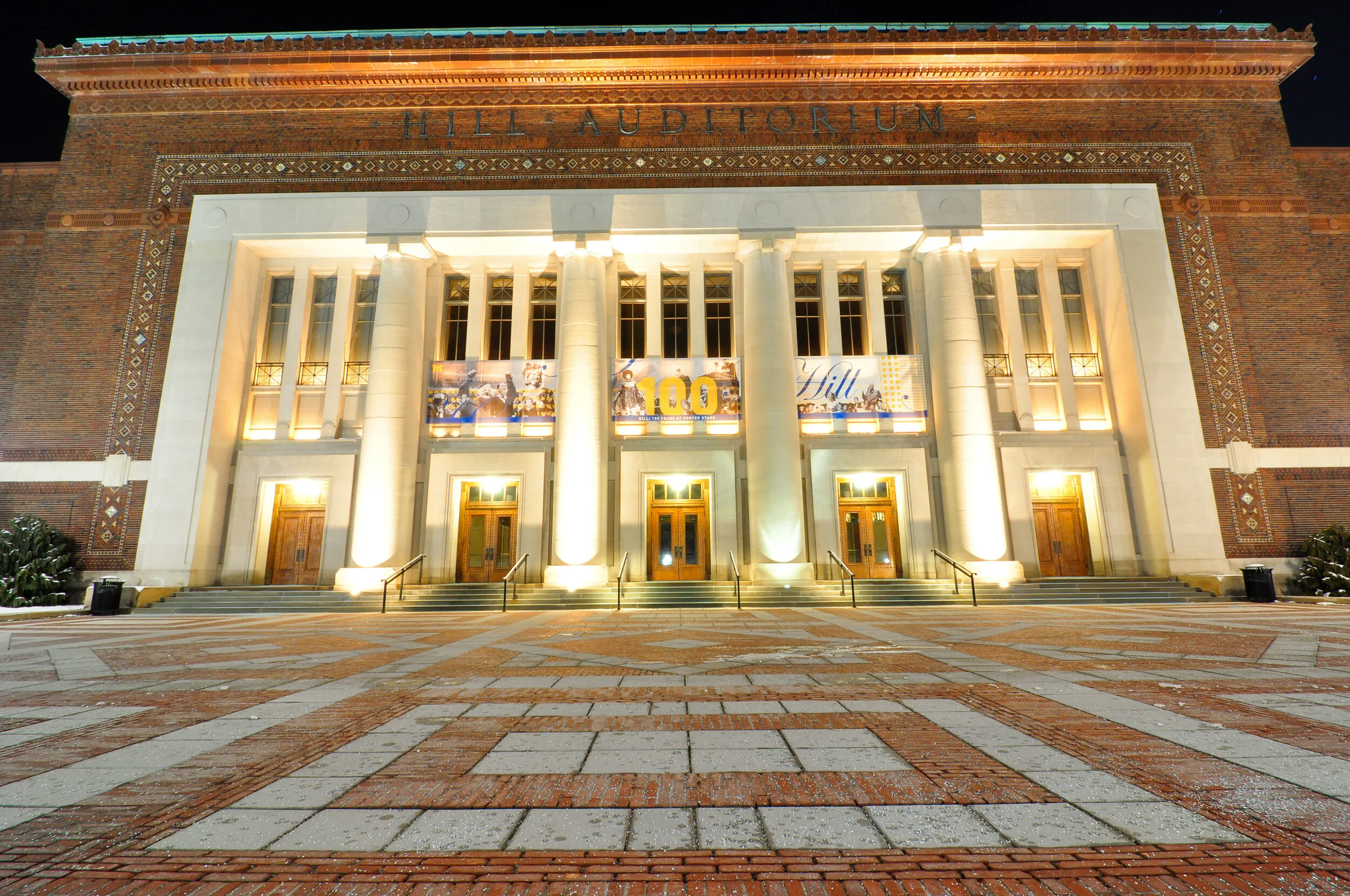 The Hill Auditorium, a large brick building with white columns and illuminated doors, decorated with banners celebrating 100 years, at night with a patterned brick courtyard in the foreground.