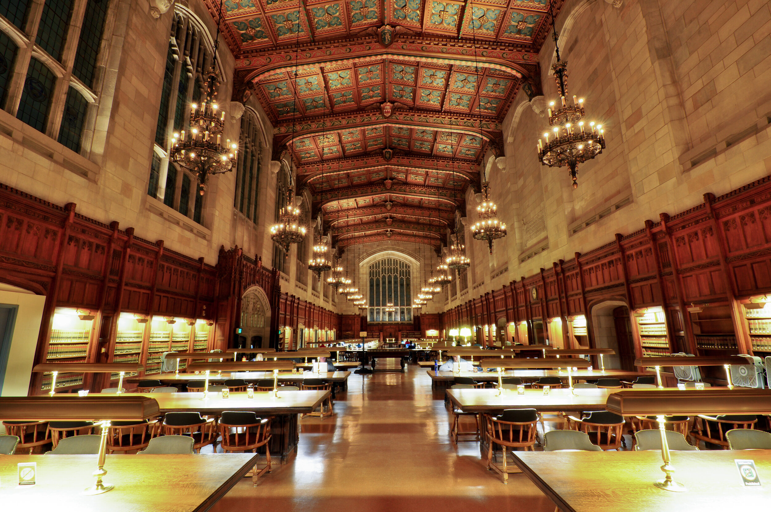 A large, historic library reading room with high vaulted ceiling, wooden paneling, chandeliers, tall windows, and long tables with lamps.
