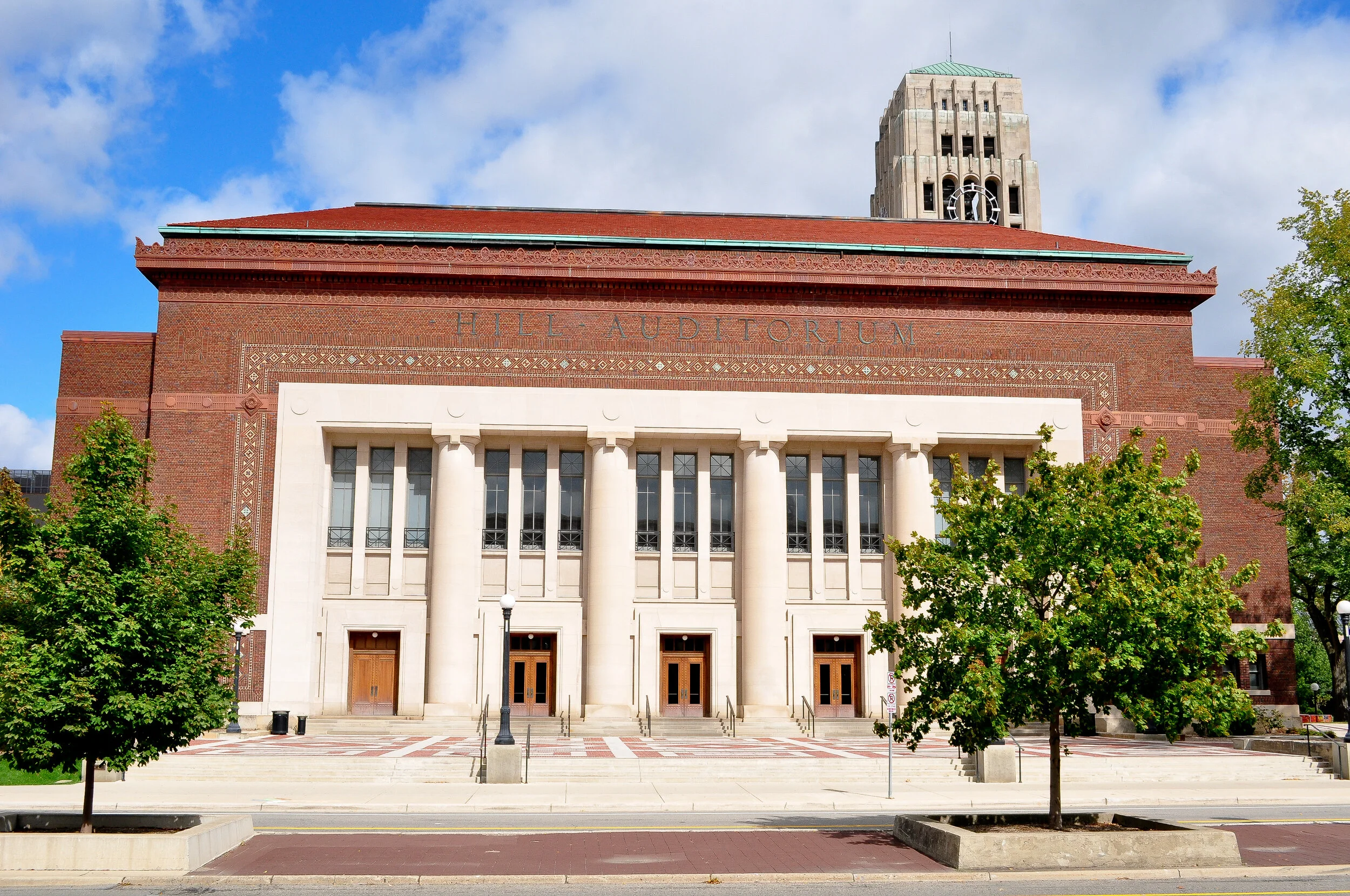 Front view of Hill Auditorium, a large brick building with white columns and multiple tall windows. Trees line the sidewalk and a clock tower is visible in the background. Clear blue sky with some clouds.