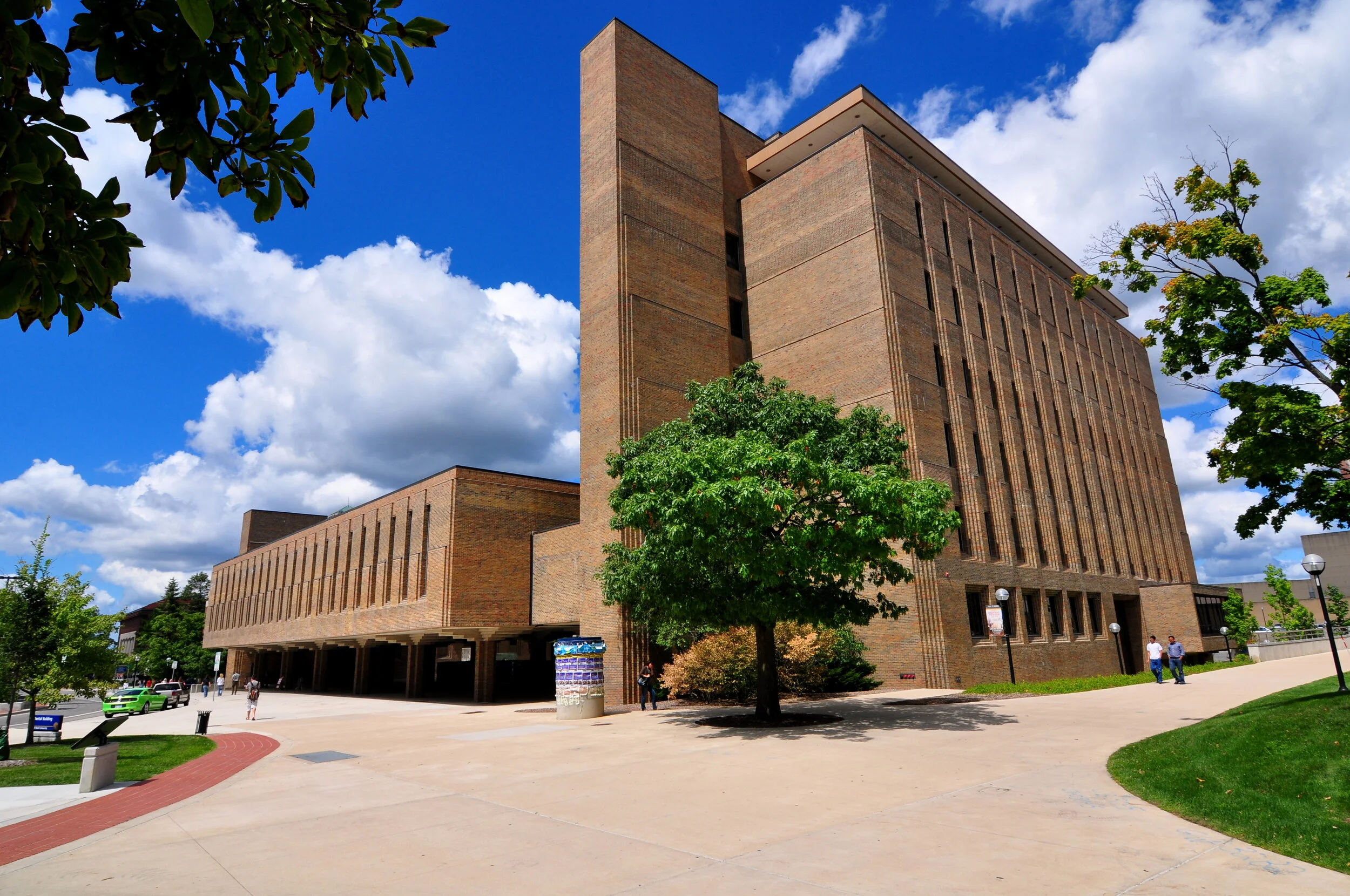 Large brick building on a university campus with trees, sidewalk, and people walking, under a partly cloudy sky.