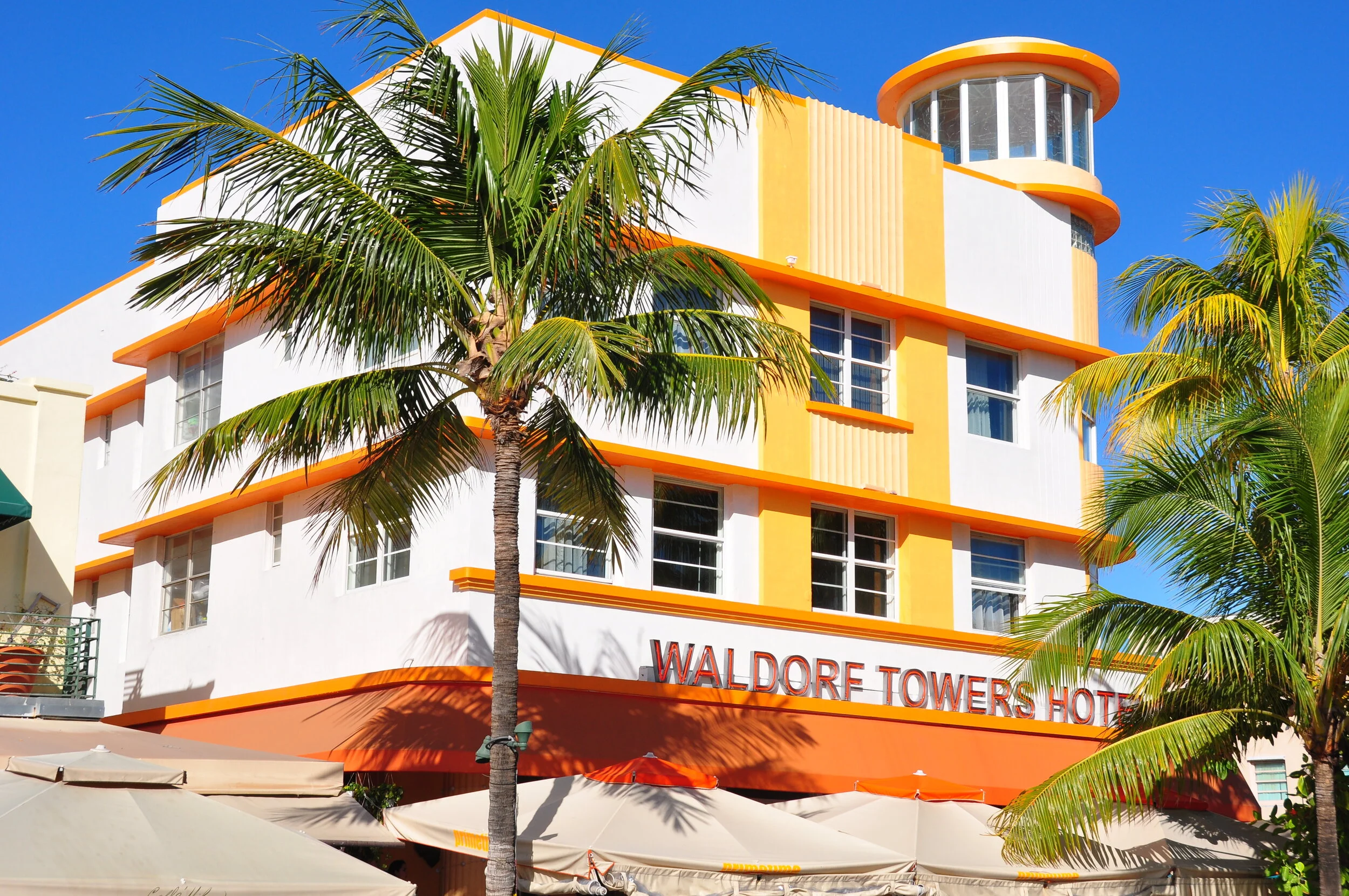 The Waldorf Towers Hotel, a white building with orange accents, surrounded by palm trees and umbrellas, against a clear blue sky.