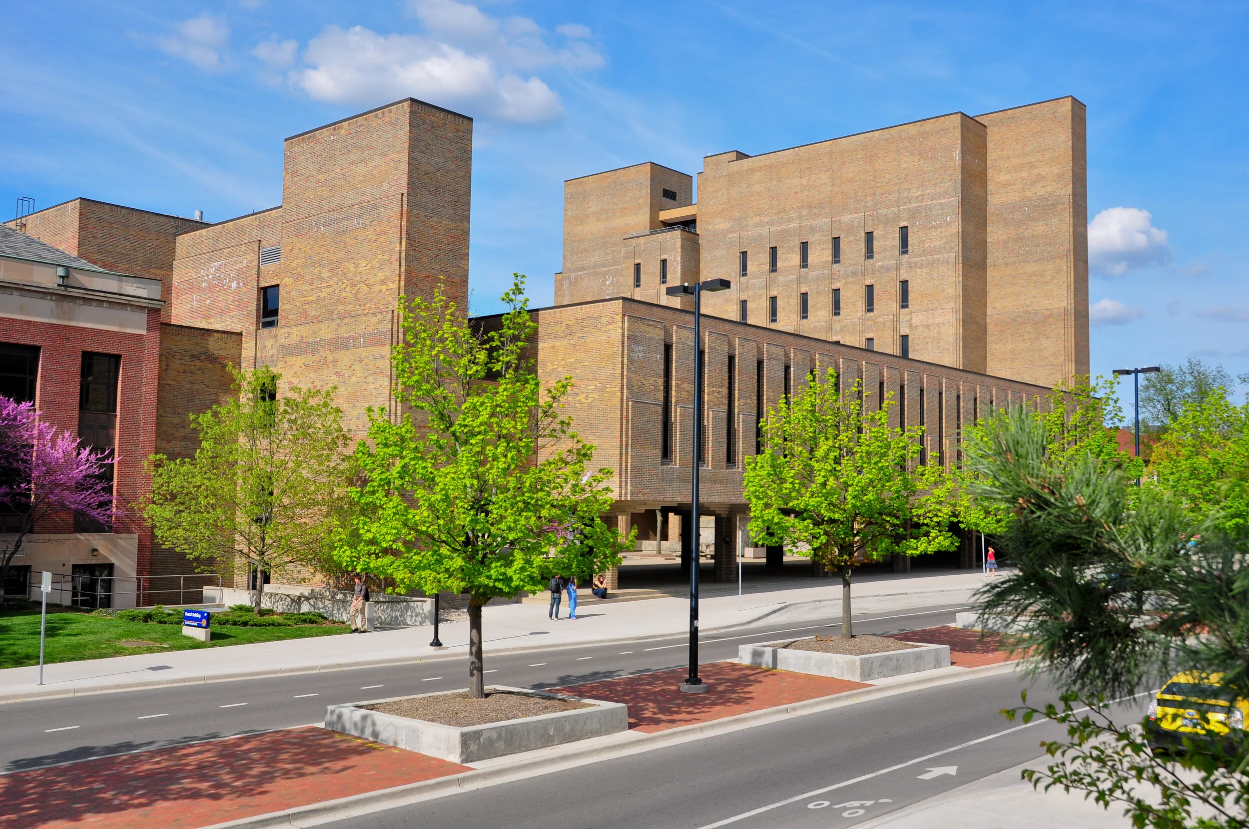 Street view of a large brick building with a modern architectural design, surrounded by trees and sidewalk, under a blue sky with some clouds.