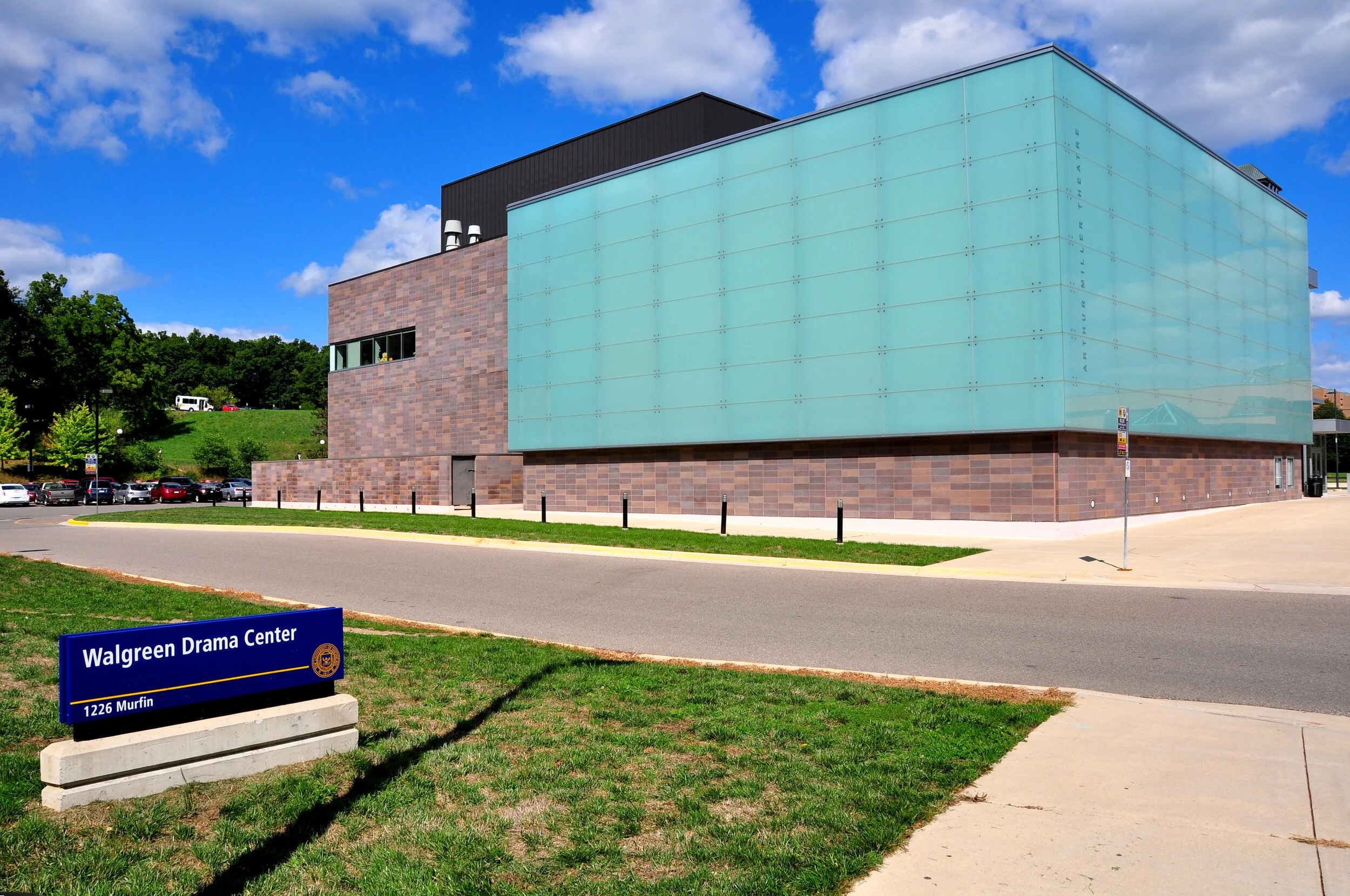 Exterior view of the Walgreens Drama Center building with a brick and glass facade, parking lot in the background, and a blue sky with clouds.