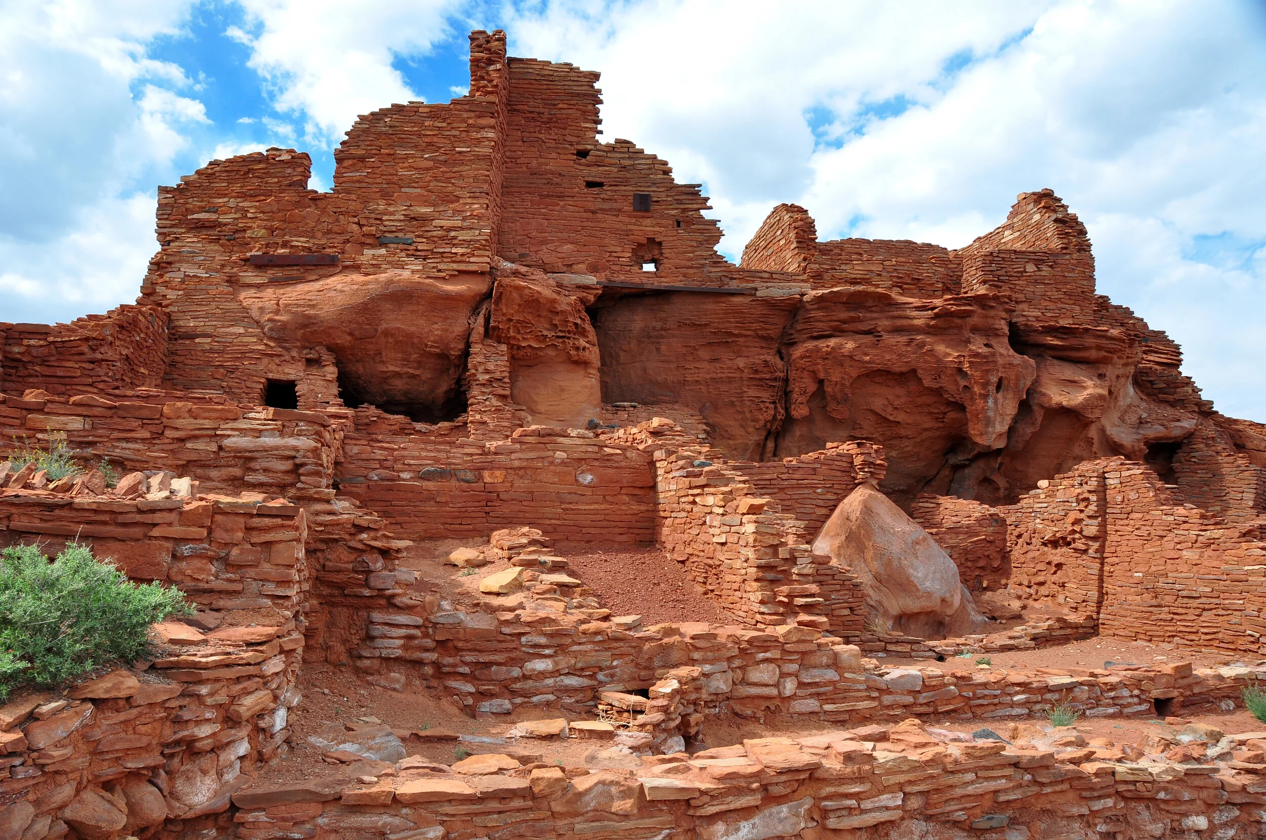Ancient red sandstone cliff dwelling ruins with small door openings under a partly cloudy sky.