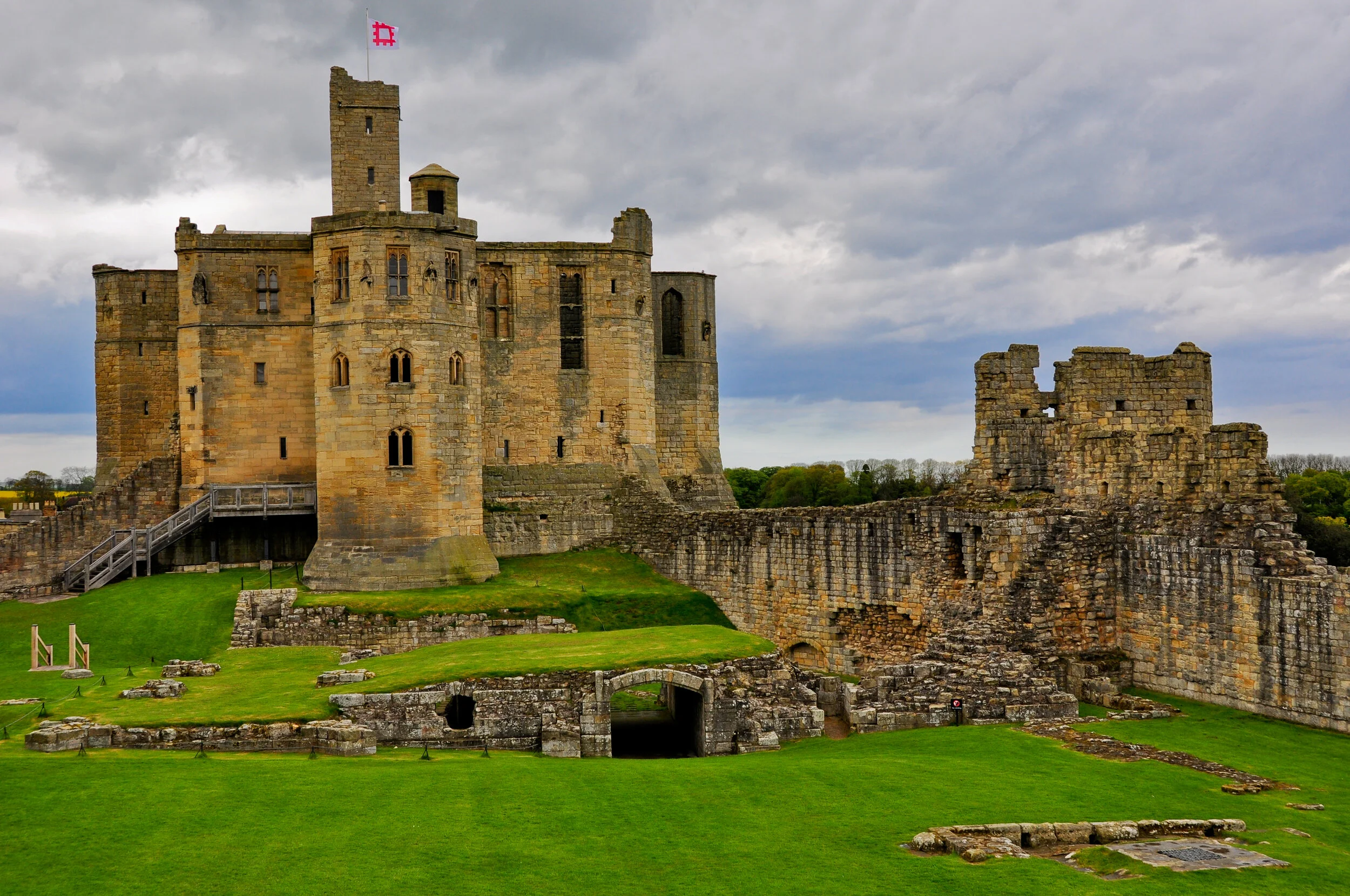 View of a historic stone castle with a flag on top, surrounded by green lawns and stone ruins under a cloudy sky.