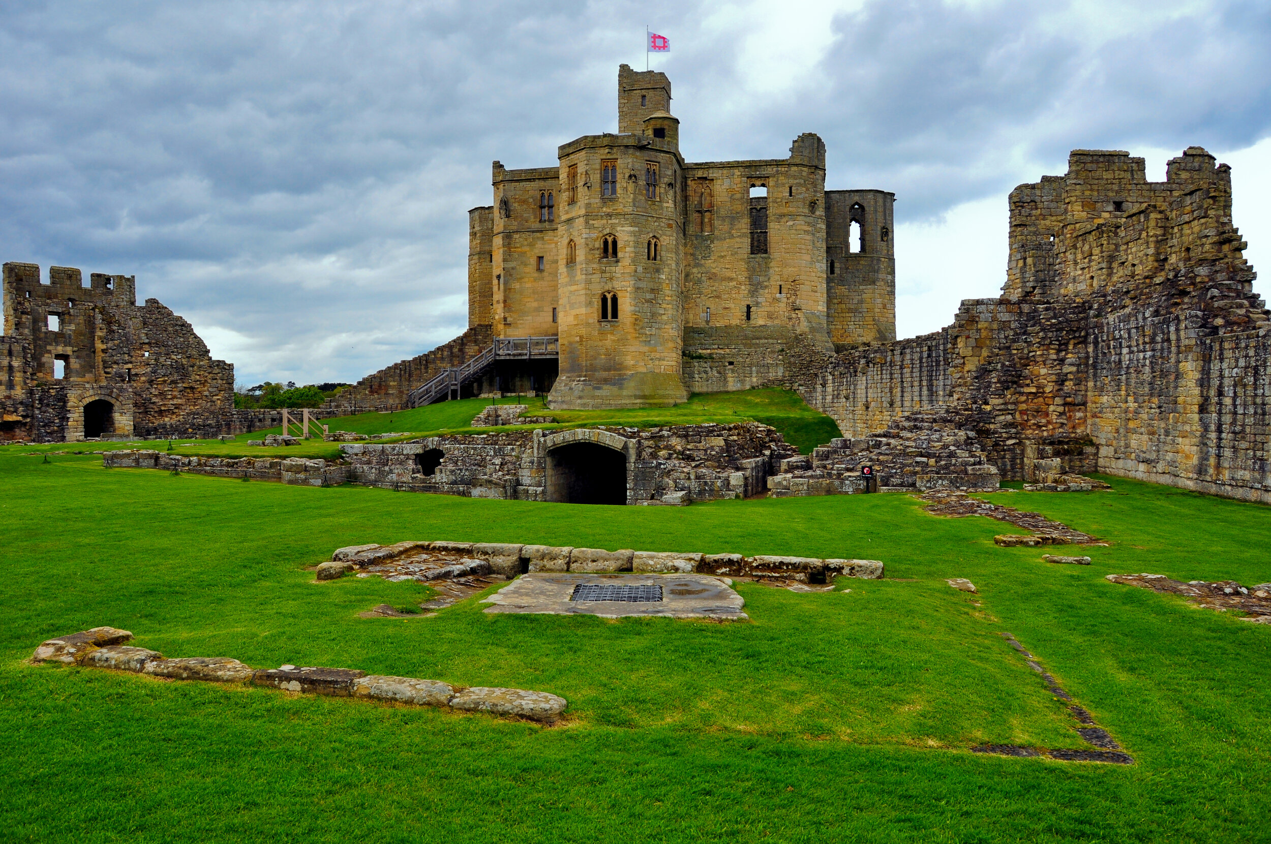 A medieval stone castle with a central tower and surrounding walls, situated on a grassy landscape under a cloudy sky.