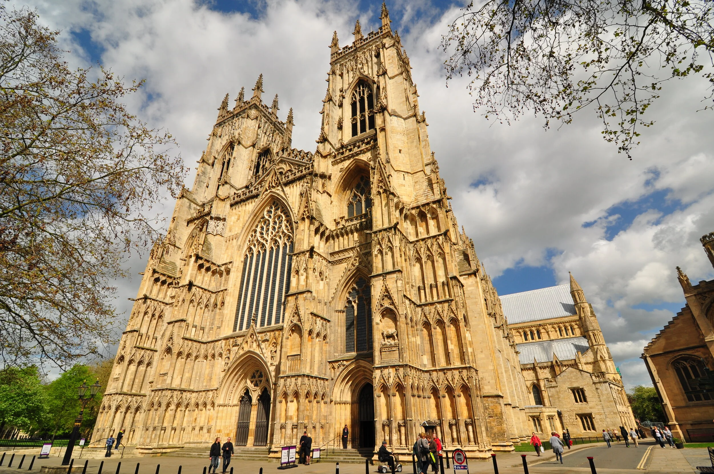 Exterior view of York Minster, a Gothic cathedral in York, England, with people walking nearby and partly cloudy sky above.