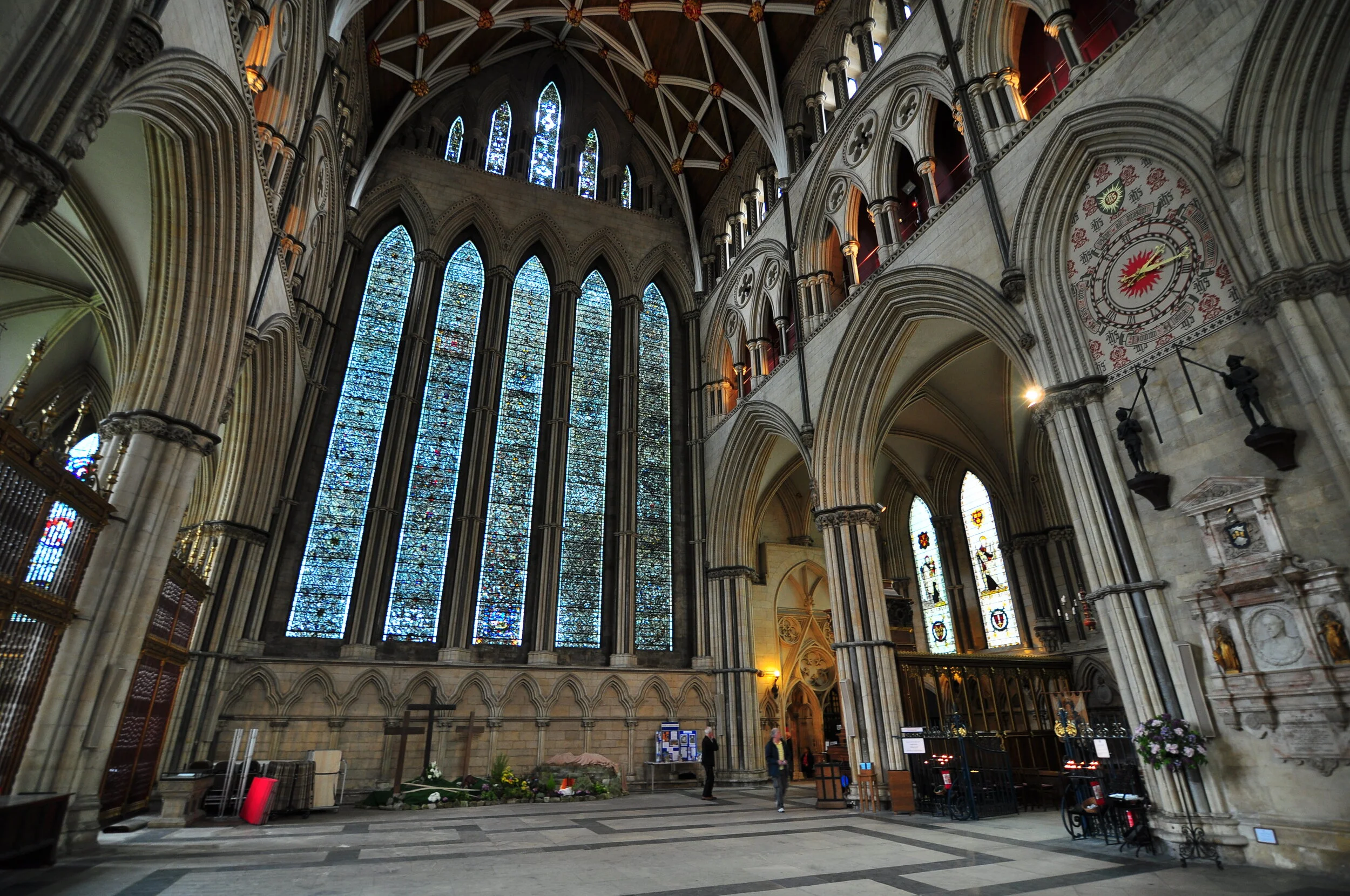 Interior of a Gothic cathedral featuring large stained glass windows, high vaulted ceilings, and stone arches. There are statues and ornate architectural details along the walls, and a few people walking inside.