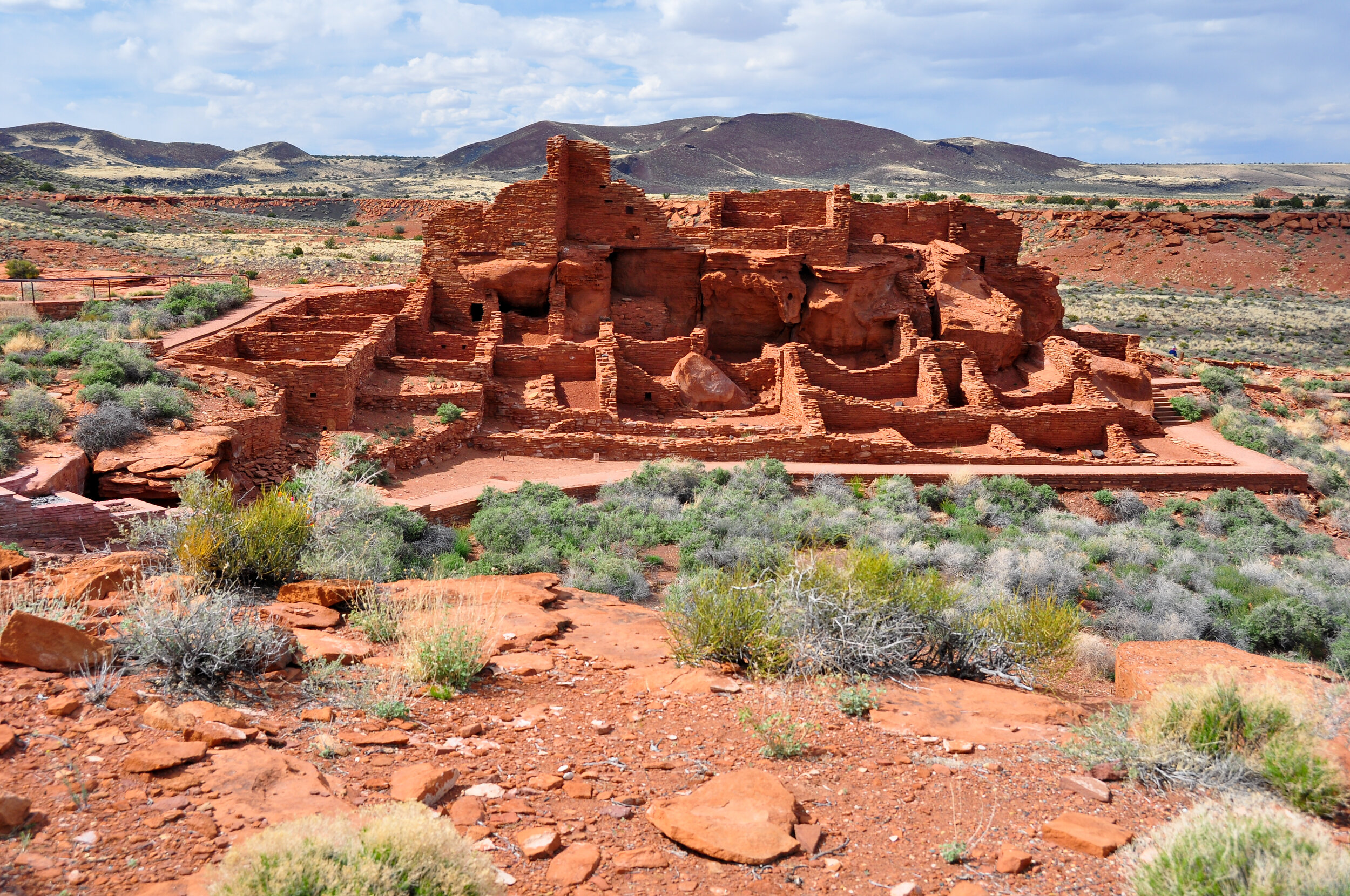 Ancient red sandstone cliff dwellings in a desert landscape with sparse vegetation, rolling hills, and a partly cloudy sky.
