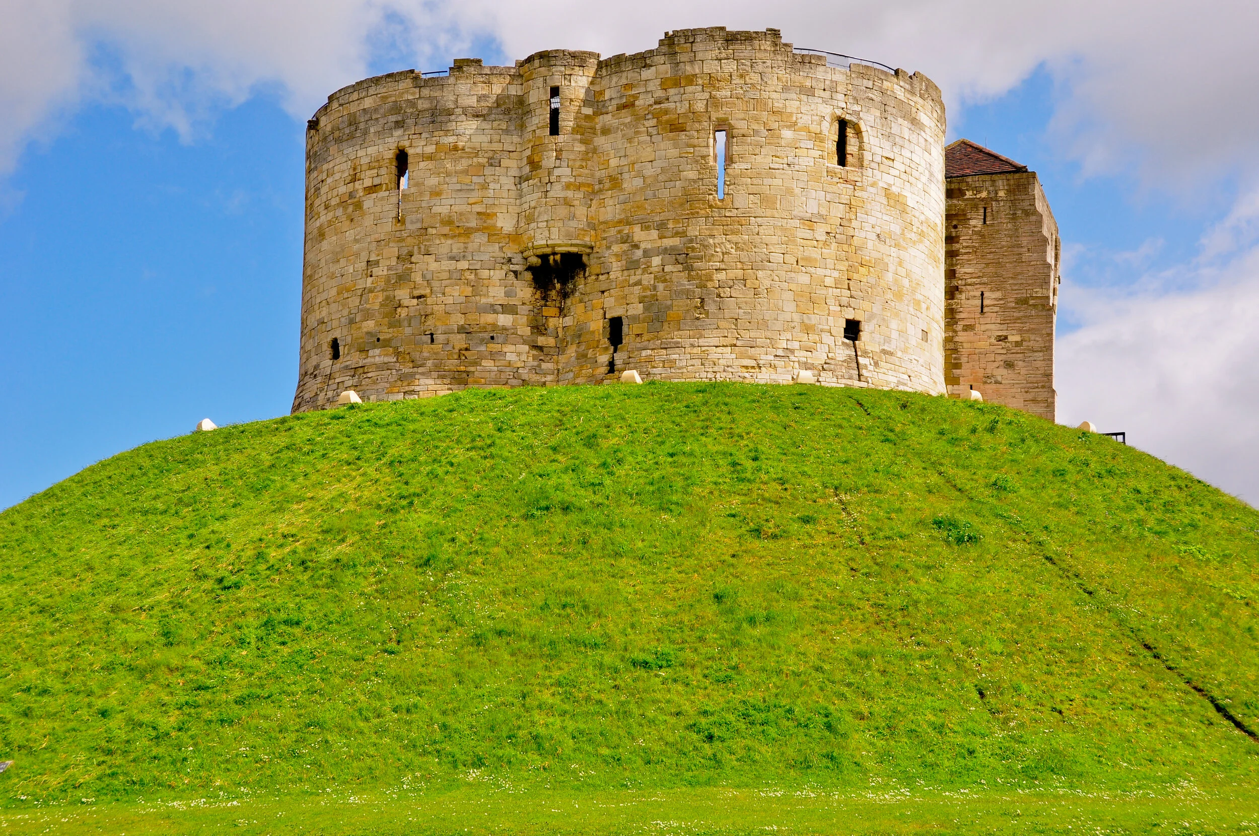 A historic stone castle on top of a grassy hill, with a blue sky and some clouds in the background.