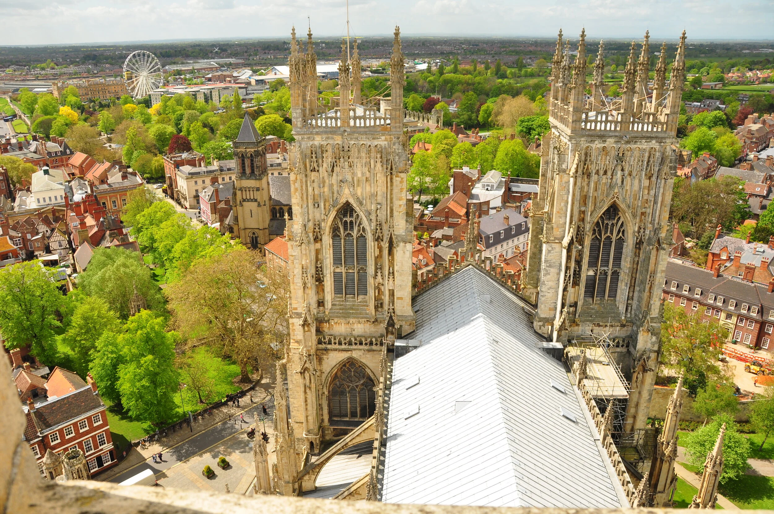 Aerial view of a Gothic cathedral with two tall spires, surrounded by city rooftops, trees, and a town in the background.