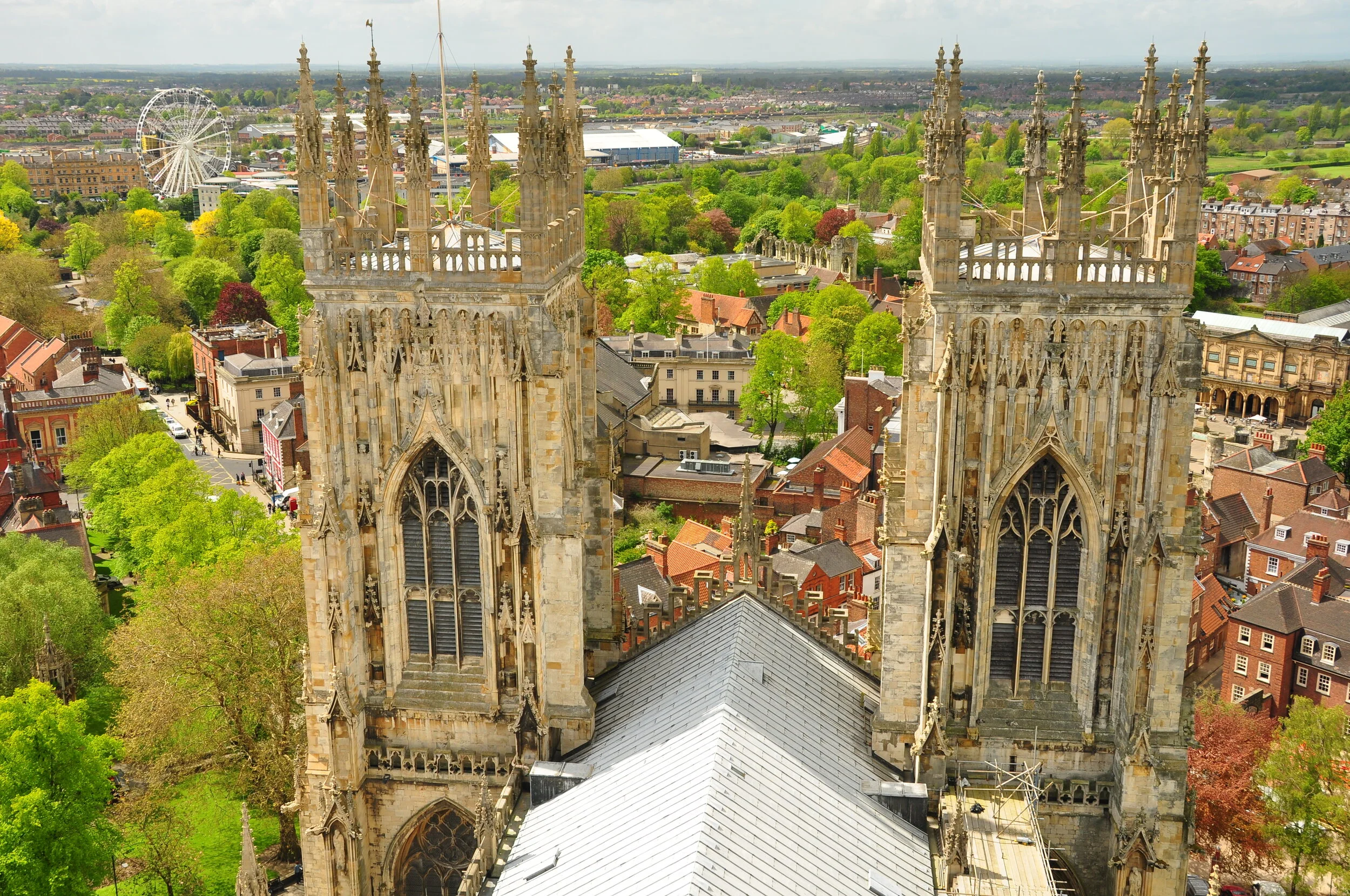 Aerial view of a historic Gothic-style cathedral with two prominent towers, surrounded by green trees and a cityscape with a Ferris wheel in the background.