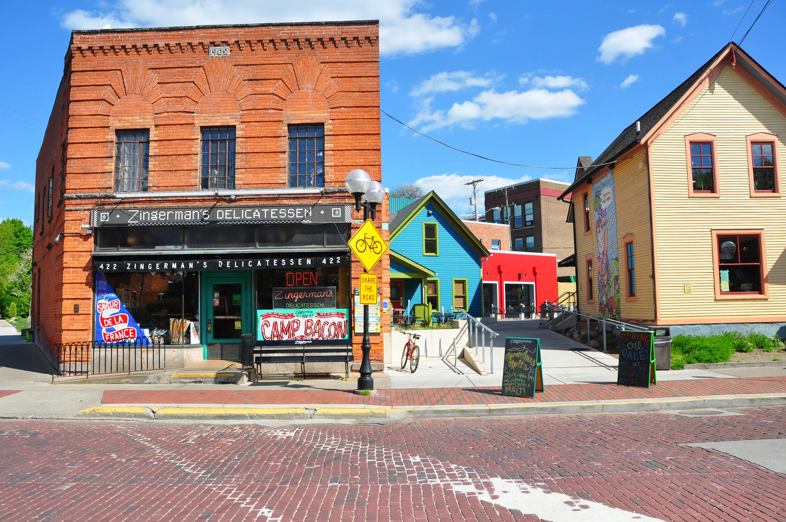 Colorful storefronts on a sunny day, including a brick building with a deli, a bright yellow house, a red building with a mural, and a blue house, with sidewalk signs, a bicycle, and a street with cobblestones.