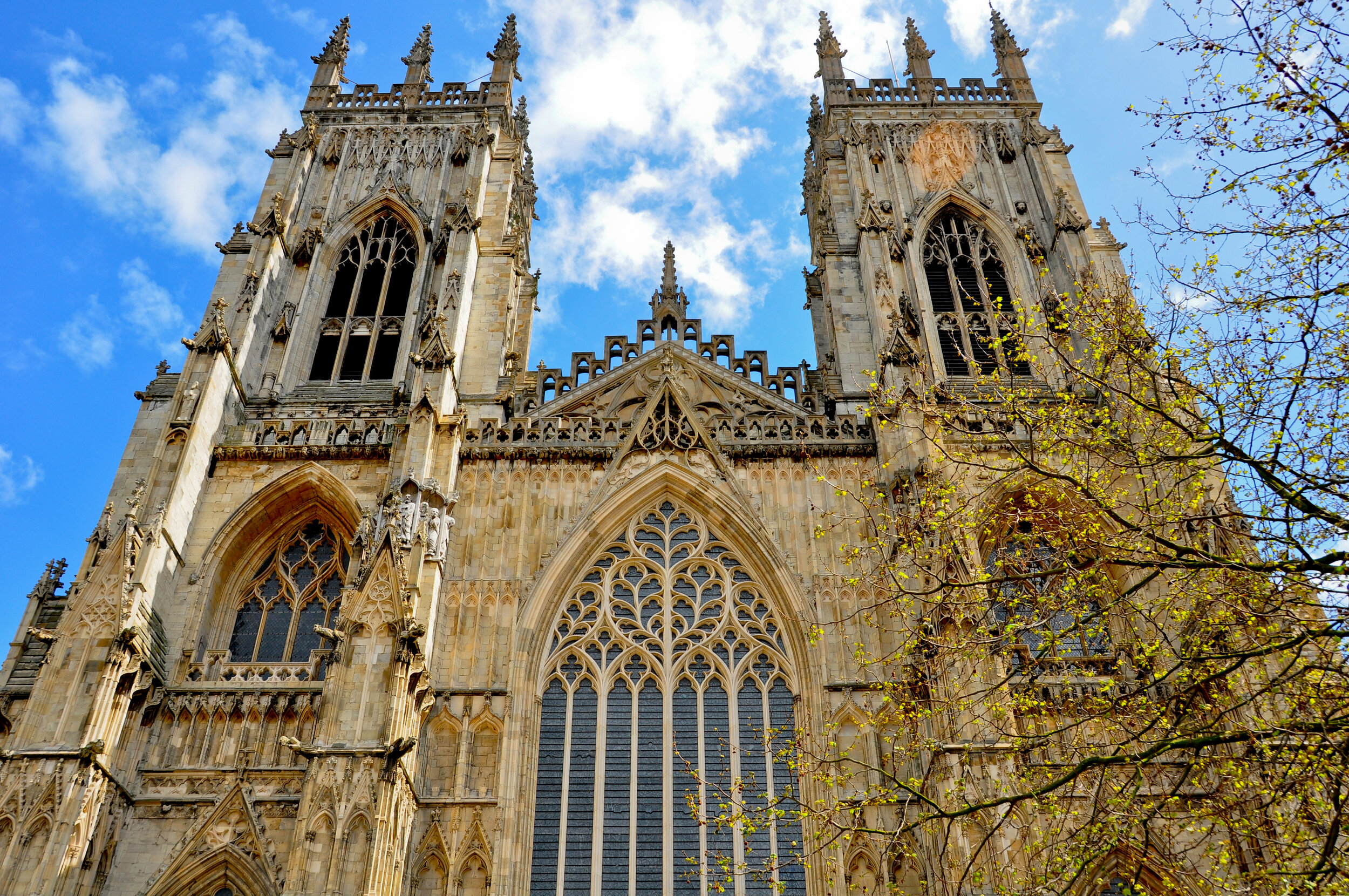 Front view of a Gothic cathedral with two tall spires, large ornate stained glass windows, and intricate stone carvings, with some tree branches in the foreground and a blue sky with clouds in the background.