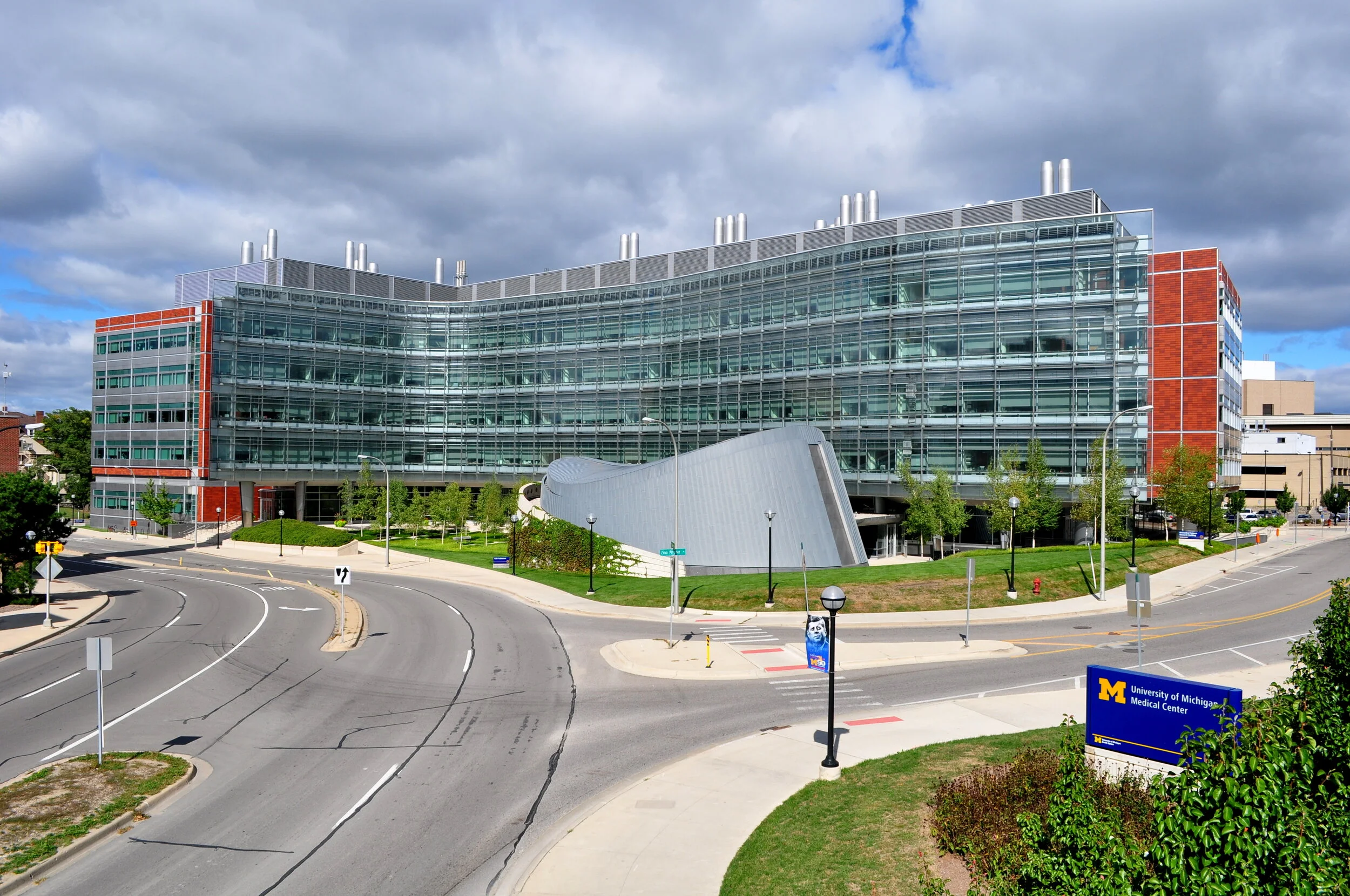 Modern medical building with glass windows, a curved metallic structure, trees, streetlights, and a blue sign that reads 'University of Michigan Medical Center' on a partly cloudy day.