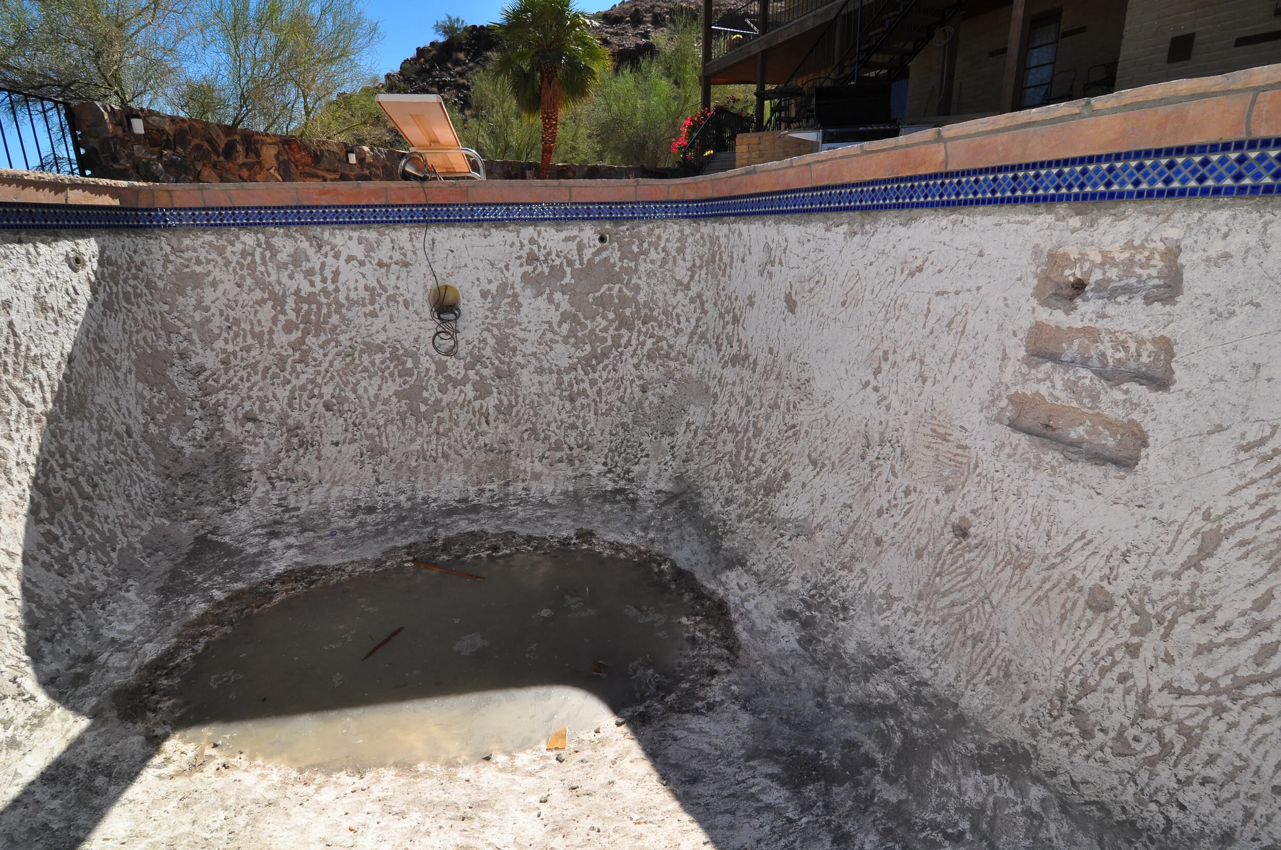 Empty swimming pool with dirty water and debris, partially shaded, with a blue tiled border on top, in a backyard with desert vegetation and mountains in the background.