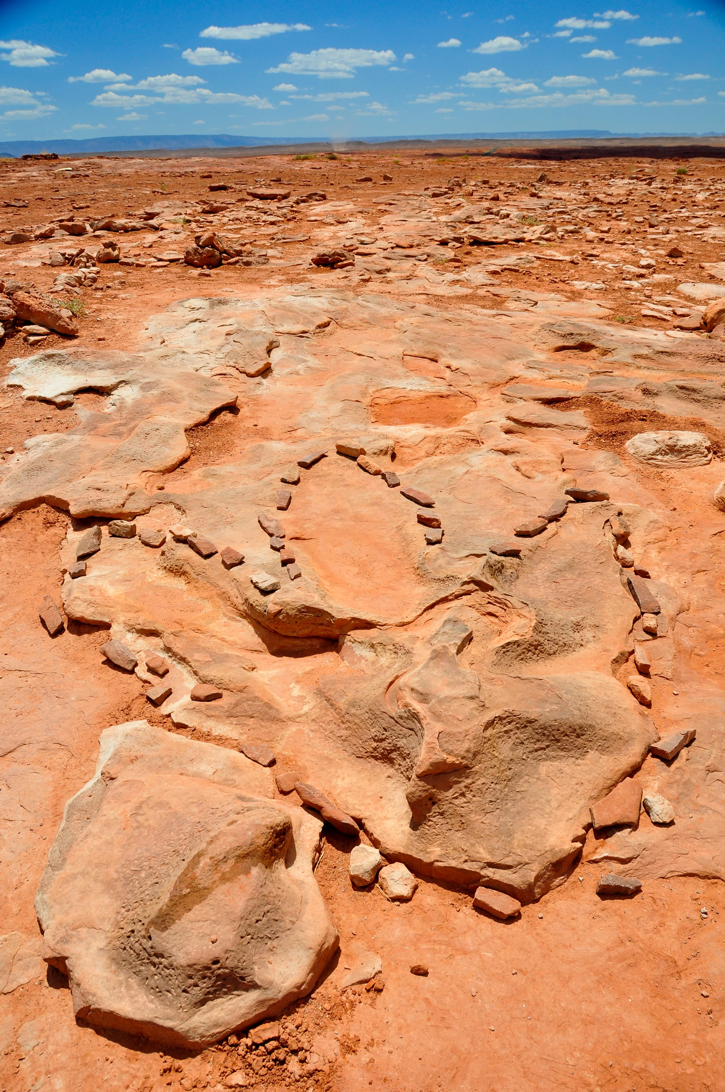 Desert landscape with a small rock formation outlined by smaller rocks, under a partly cloudy blue sky.