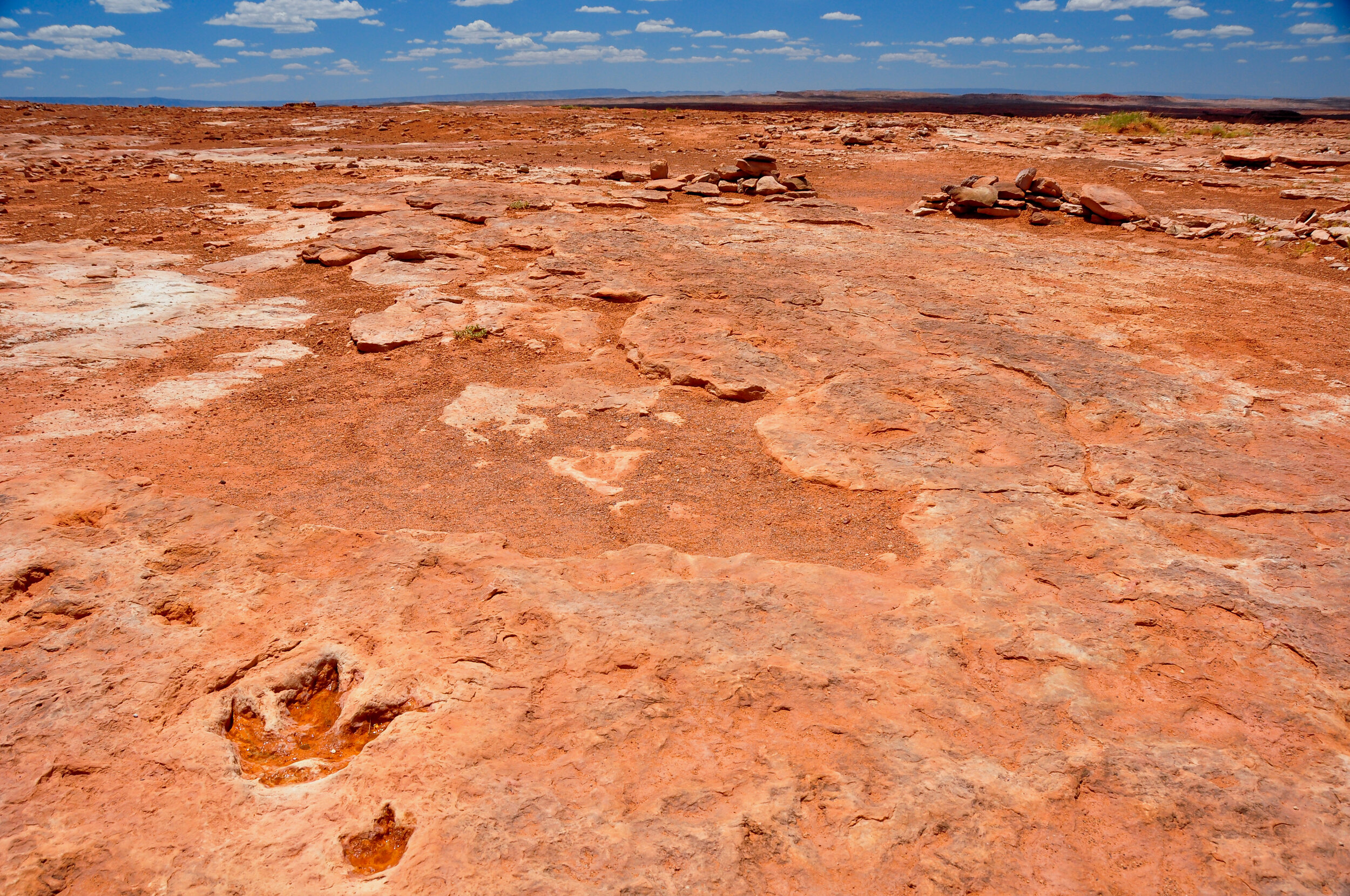 A vast, flat desert landscape with reddish-brown rocky terrain under a partly cloudy blue sky.