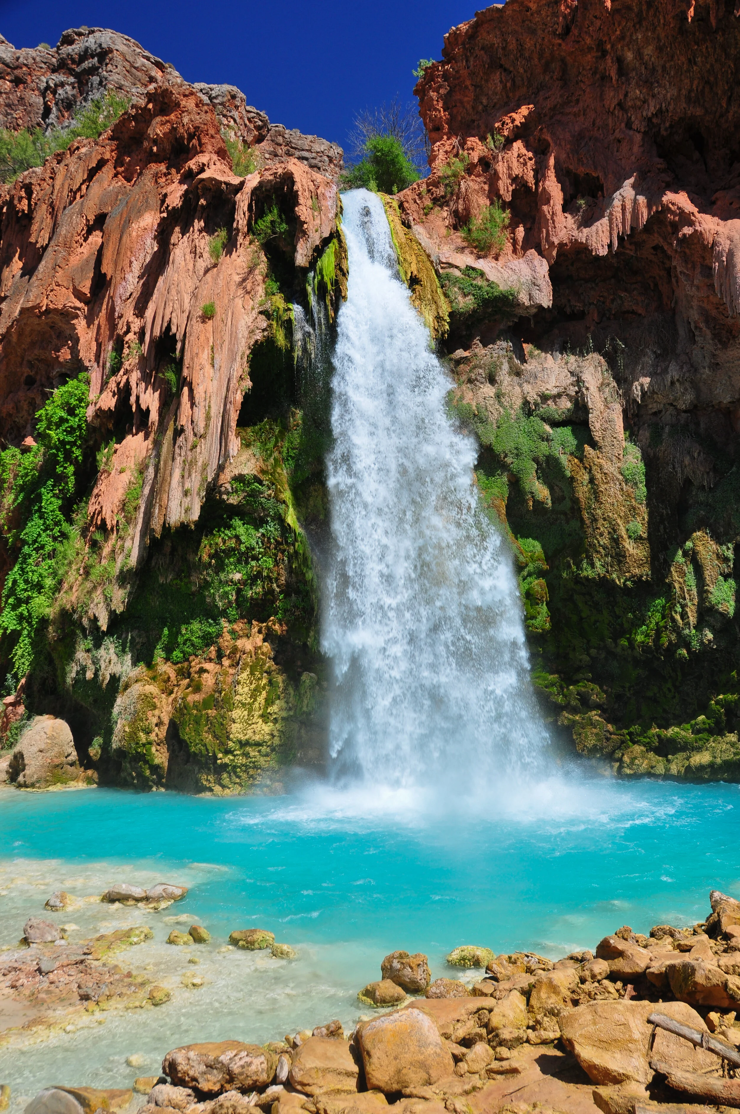 A waterfall cascading into a blue pool surrounded by rocks and red rock cliffs, with green vegetation and a clear blue sky above.