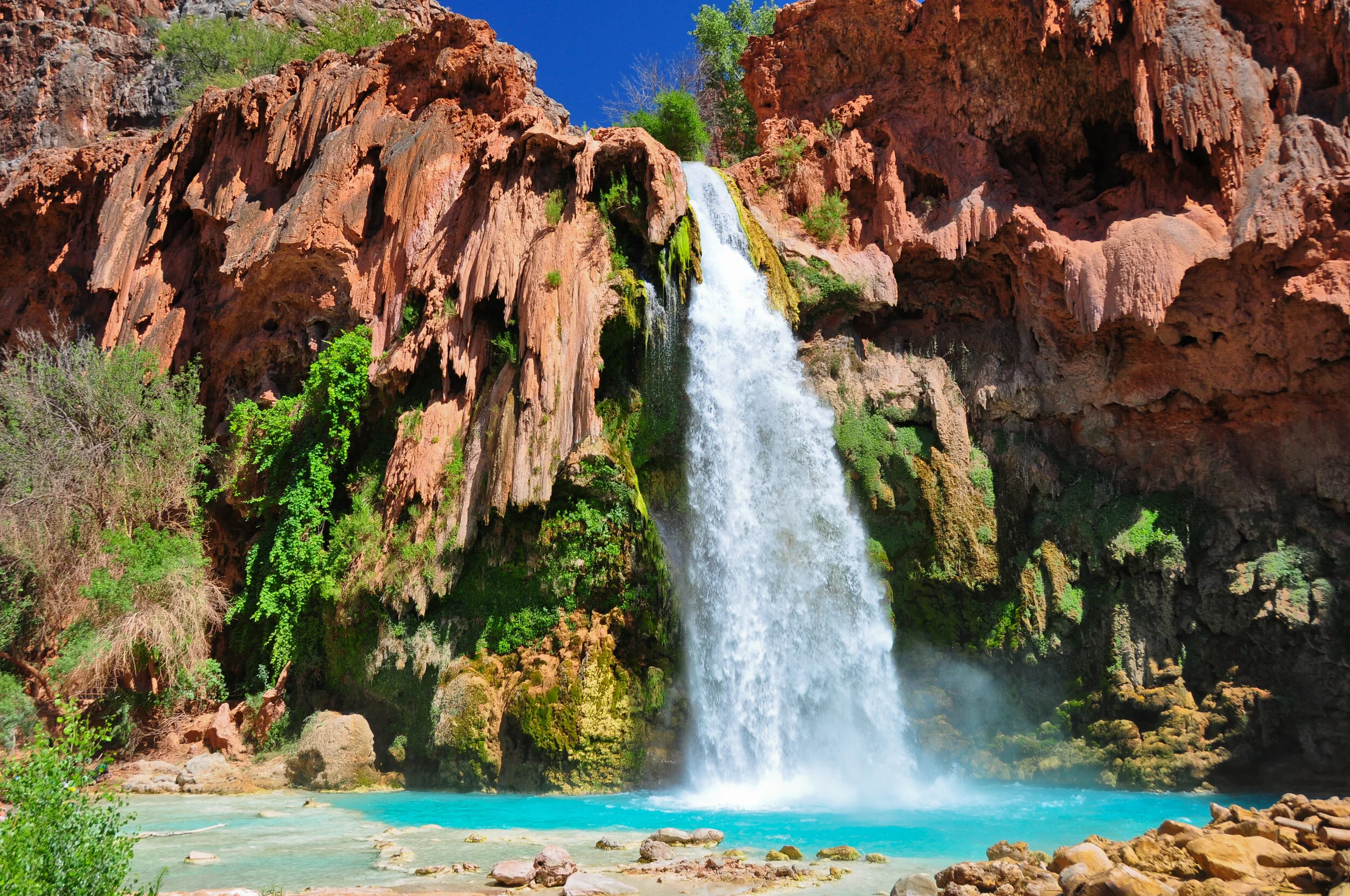 A waterfall cascading from a red rocky cliff into a turquoise pool surrounded by lush green foliage.
